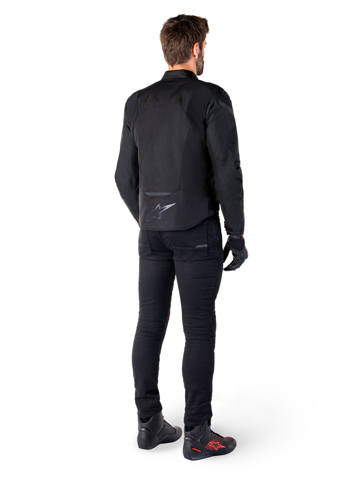 Alpinestars T-Jaws V4 Waterproof Jacket, nero, vista posteriore su modello con pantaloni e stivali da guida neri, che mette in risalto la costruzione in tessuto sportivo e il logo Alpinestars sulla parte bassa della schiena.