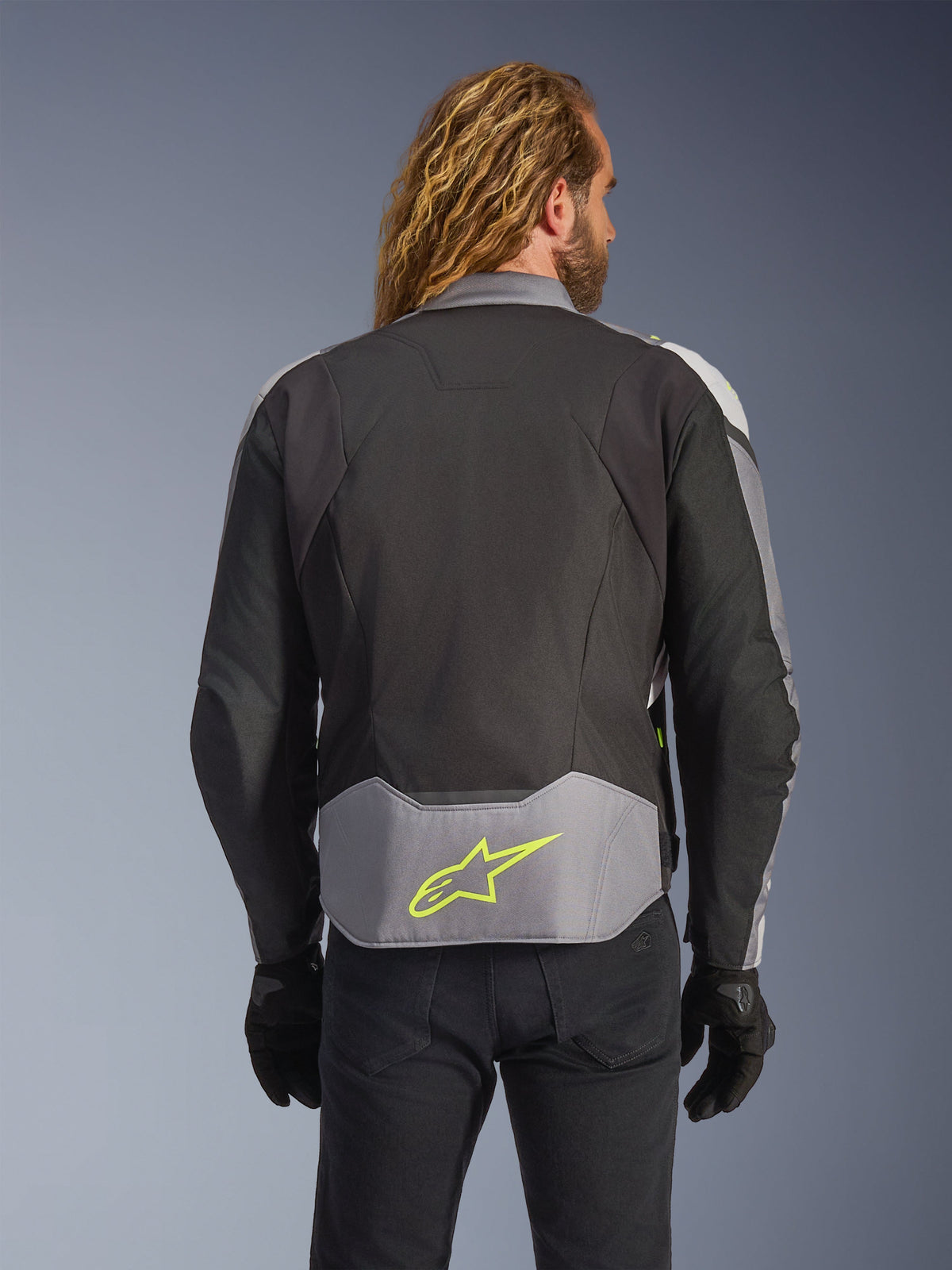 Giacca impermeabile Alpinestars T-Jaws V4, nero, grigio ghiaccio e grigio scuro, vista posteriore su modello, caratterizzata da un pannello grigio chiaro sulla parte bassa della schiena con un prominente logo a stella giallo neon, costruzione in tessuto multi-pannello e silhouette sportiva, indossata con guanti neri e pantaloni scuri.