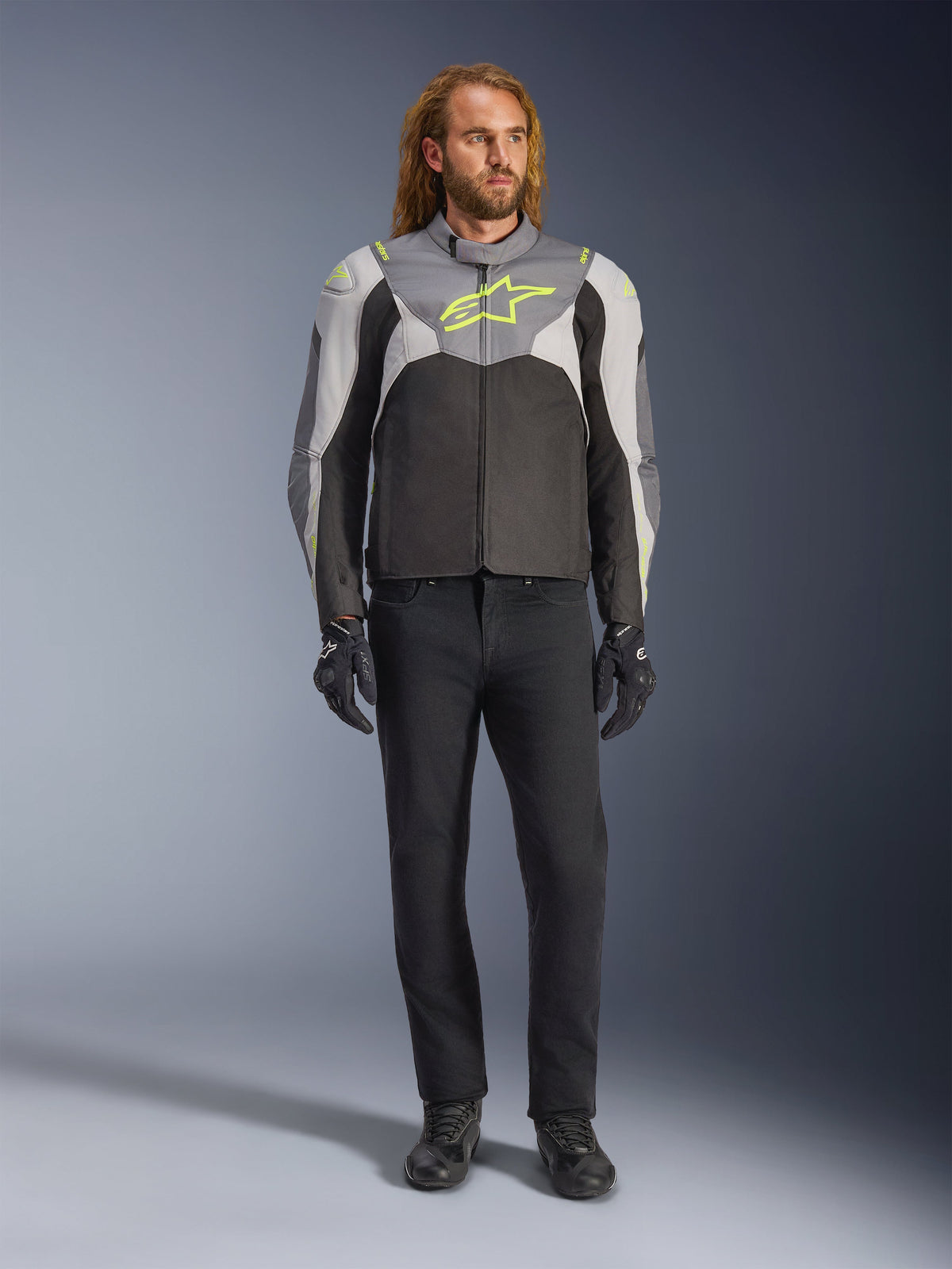 Giacca Alpinestars T-Jaws V4 Waterproof in Black Ice Gray Dark Gray con accenti giallo neon, indossata da un modello, caratterizzata da una silhouette sportiva, costruzione in tessuto tecnico e prominente logo sul petto, abbinata a pantaloni da guida e guanti neri