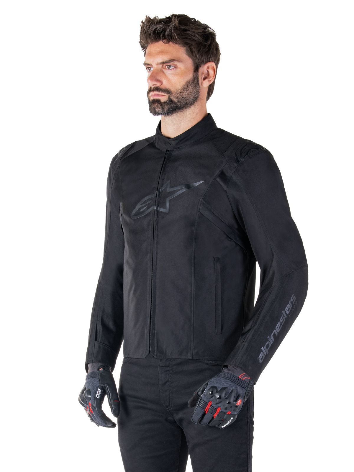 Giacca Alpinestars T-SPS V2 Waterproof, nera, indossata con pantaloni neri e guanti da moto grigi con accenti rossi, con logo nero discreto sul petto e branding sulla manica, giacca in tessuto dal taglio sportivo per guida su strada