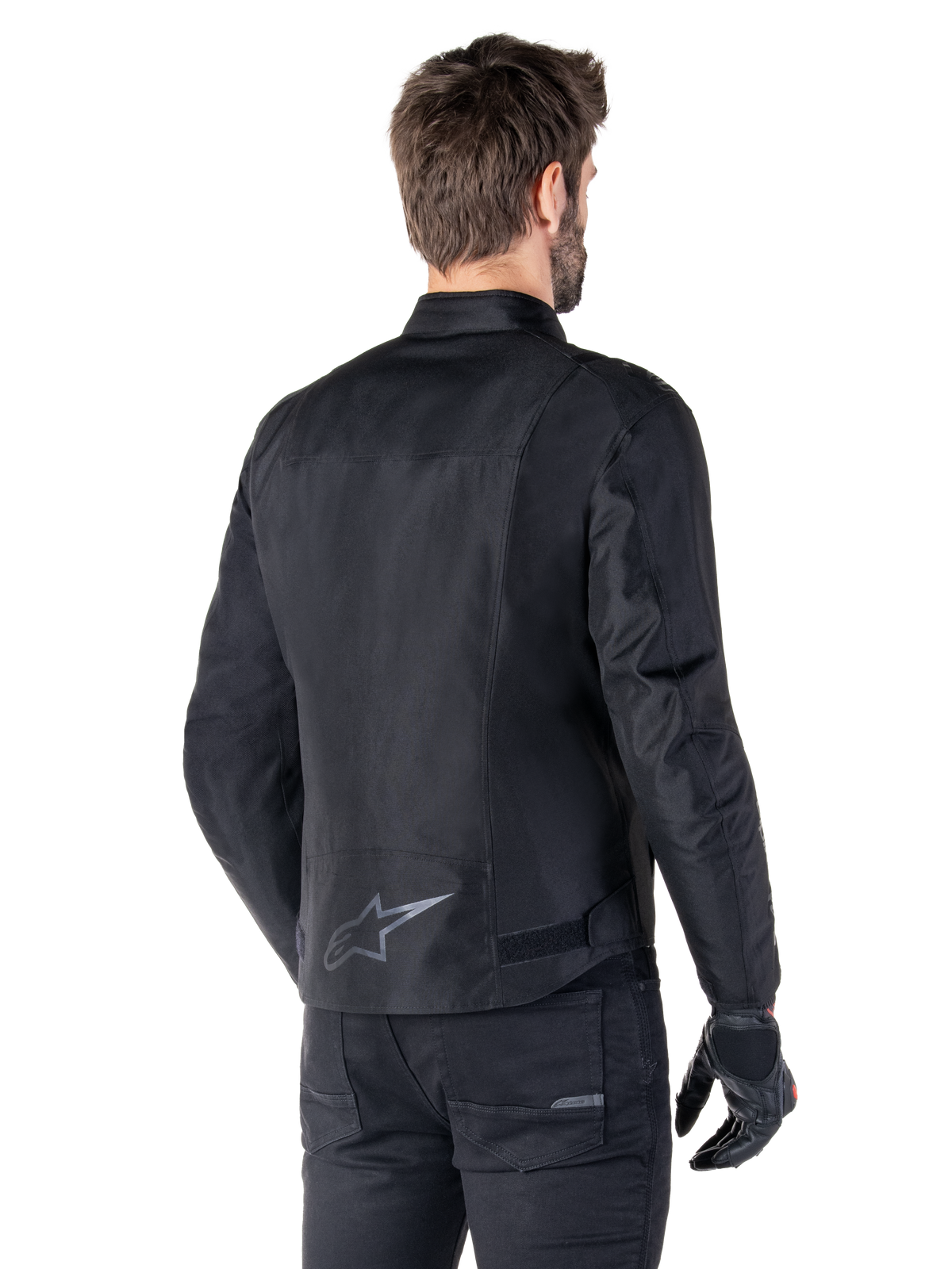 Giacca impermeabile Alpinestars T-SPS V2, nero, vista posteriore su un modello, con logo a stella riflettente sulla parte bassa della schiena, profilo posteriore allungato e cinghie di regolazione in vita, abbinata a pantaloni e guanti da moto neri