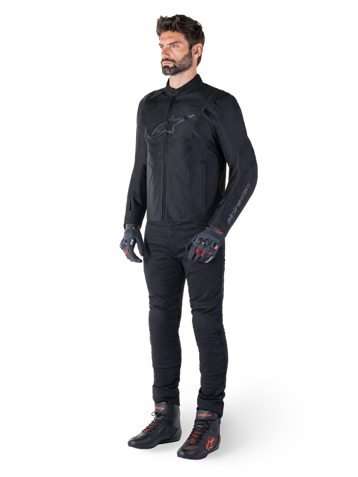 Giacca impermeabile Alpinestars T-SPS V2, nero, indossata da un modello con pantaloni neri coordinati, guanti neri con dettagli rossi e scarpe da moto nere, equipaggiamento completo da moto per guida urbana e sportiva, sfondo bianco