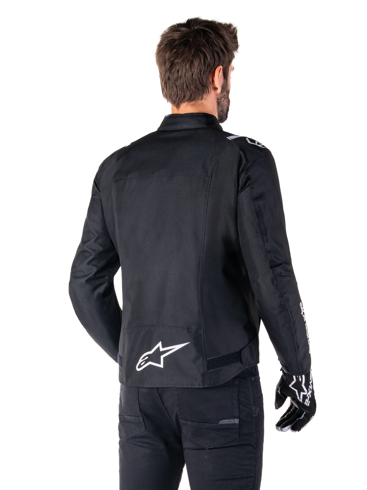 Giacca Alpinestars T-SPS V2 Waterproof, nero e bianco, vista posteriore di un motociclista che indossa la giacca, logo Alpinestars bianco sulla parte bassa della schiena, struttura in tessuto con taglio sportivo, indossata con pantaloni da moto e guanti neri