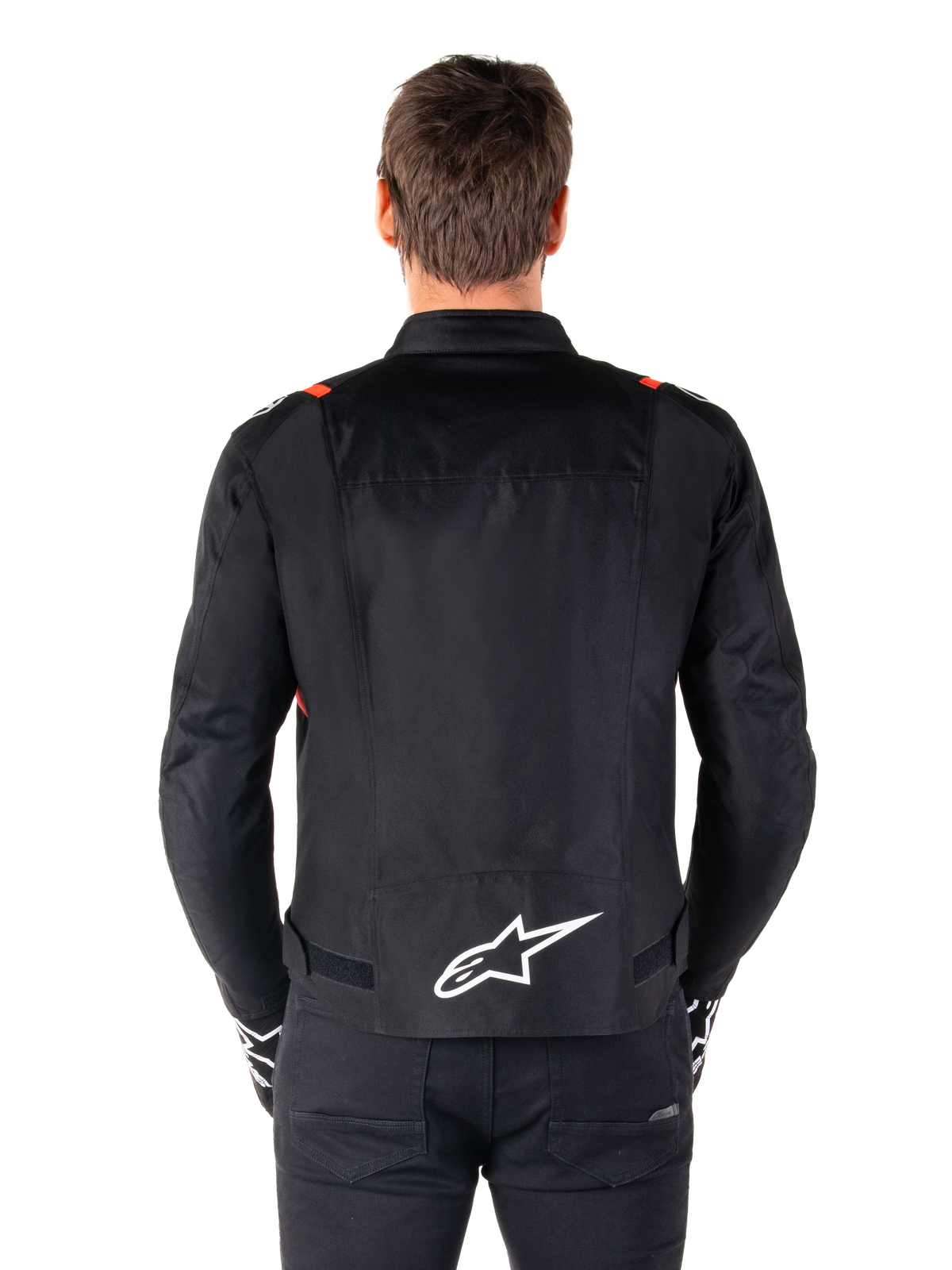 Giacca Alpinestars T-SPS V2 Waterproof, nero con accenti bianco e rosso fluo, vista posteriore con grande logo Alpinestars bianco sulla parte bassa della schiena, giacca da moto in tessuto dal taglio sportivo, indossata con pantaloni e guanti neri