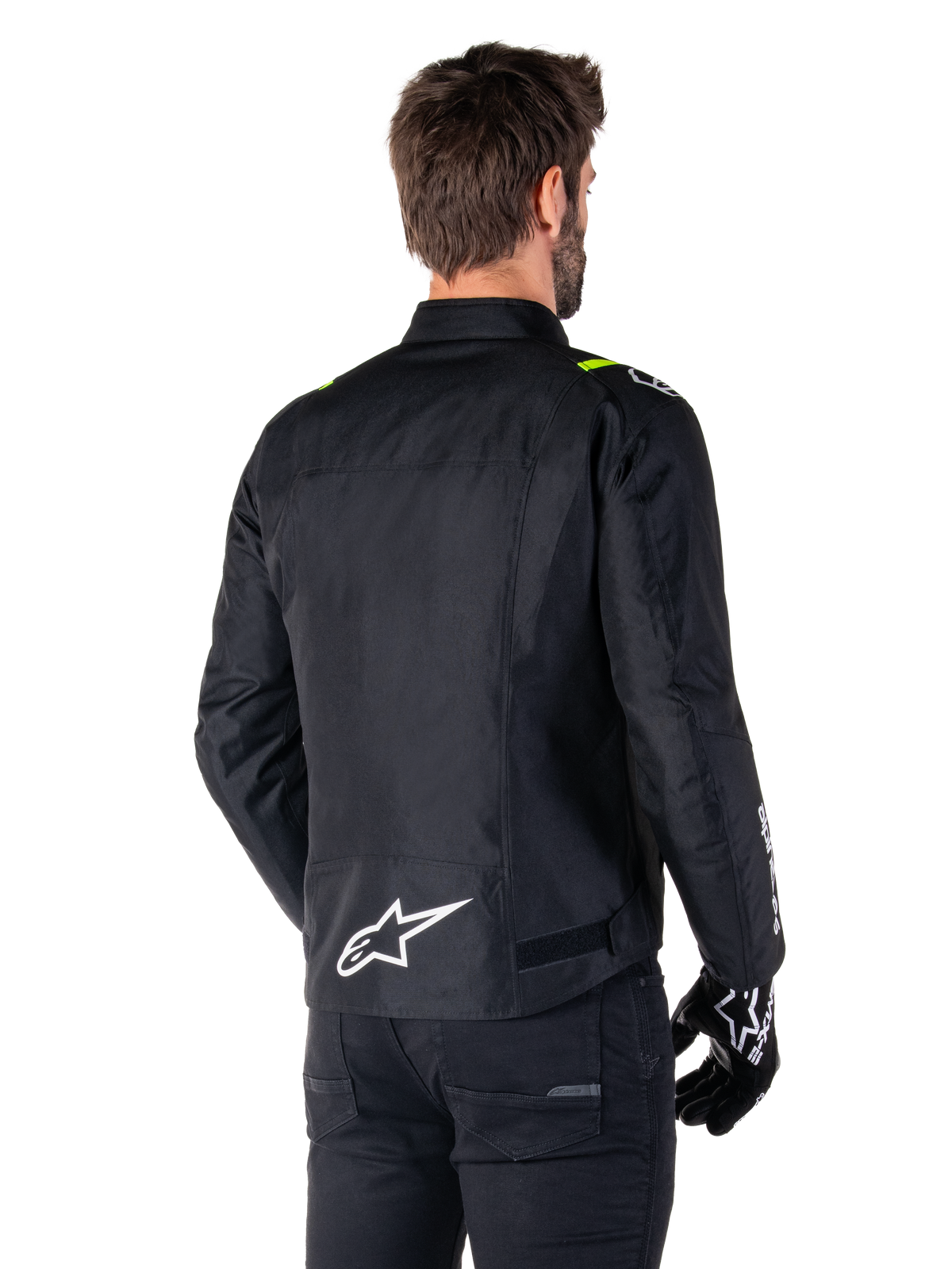 Alpinestars T-SPS V2 Waterproof Jacket, nero bianco giallo fluo, vista posteriore di un modello maschile che indossa la giacca da moto in tessuto, con logo bianco sulla parte bassa della schiena e accenti giallo fluo sulle spalle, abbinata a pantaloni e guanti neri.