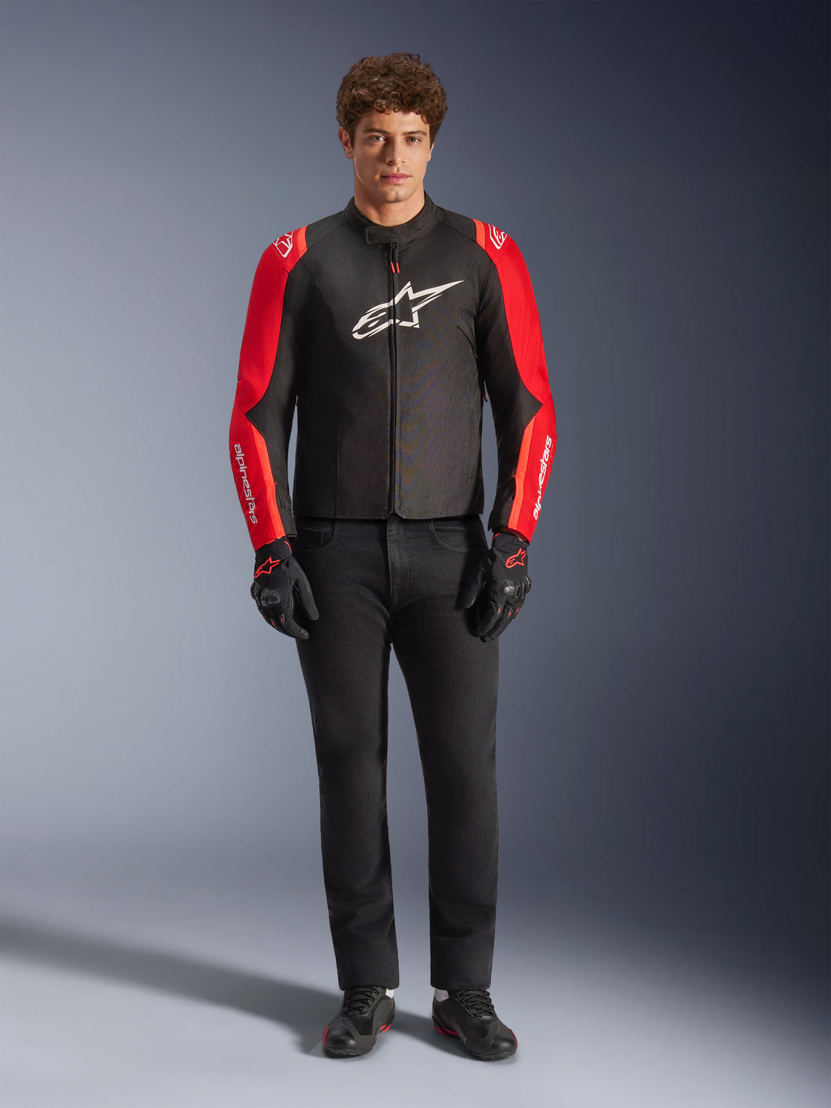 Modello che indossa la giacca Alpinestars T-SPS V2 Waterproof, nero con maniche rosse e rosso fluo, logo bianco sul petto, abbinata a pantaloni neri, guanti e scarpe da moto neri, design sportivo per abbigliamento moto stradale