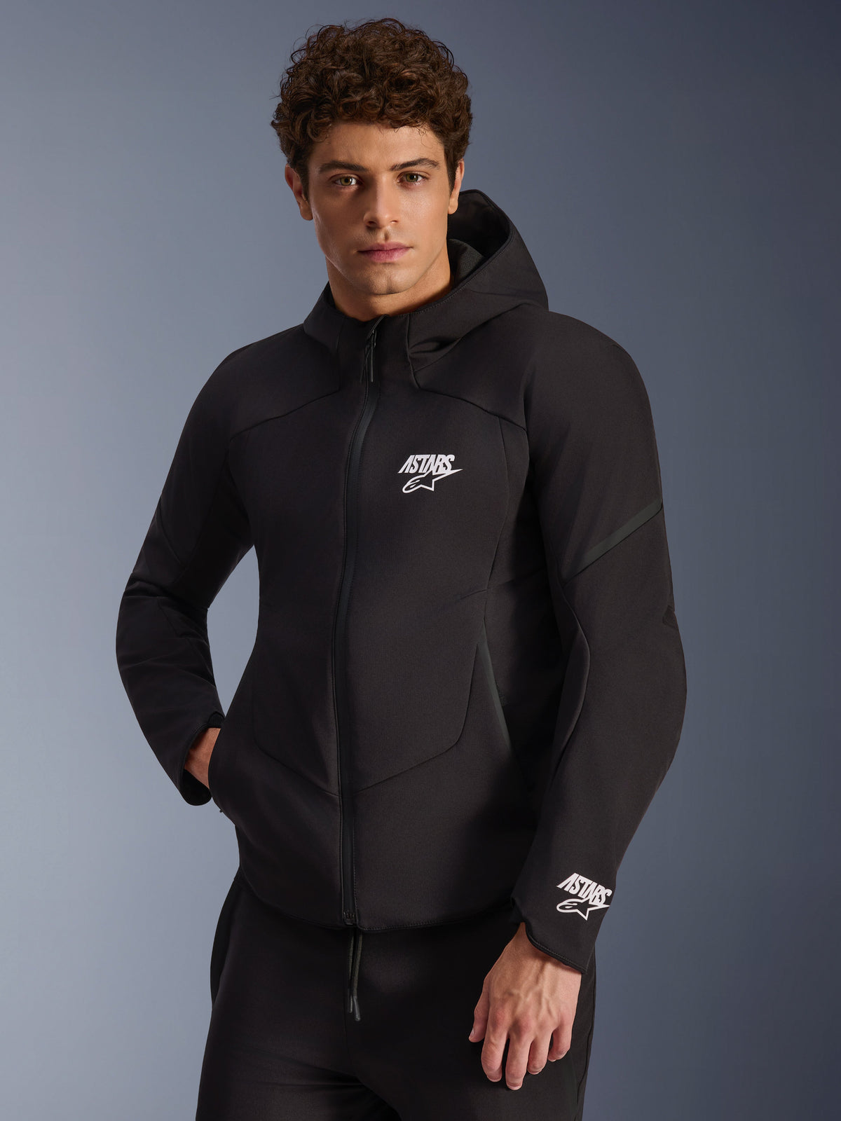 Alpinestars giacca Aeroshell impermeabile, giacca da moto softshell da uomo, nera, con loghi ASTARS bianchi e cappuccio integrato, indossata da un modello.