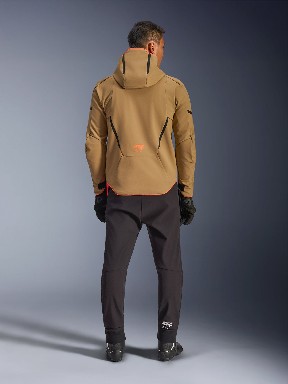 Alpinestars Aeroshell Waterproof Jacket in Dark Khaki Black Orange Fluo, vista posteriore di un uomo che indossa la giacca softshell con cappuccio, con profili neri e logo arancione, abbinata a pantaloni tecnici e guanti neri
