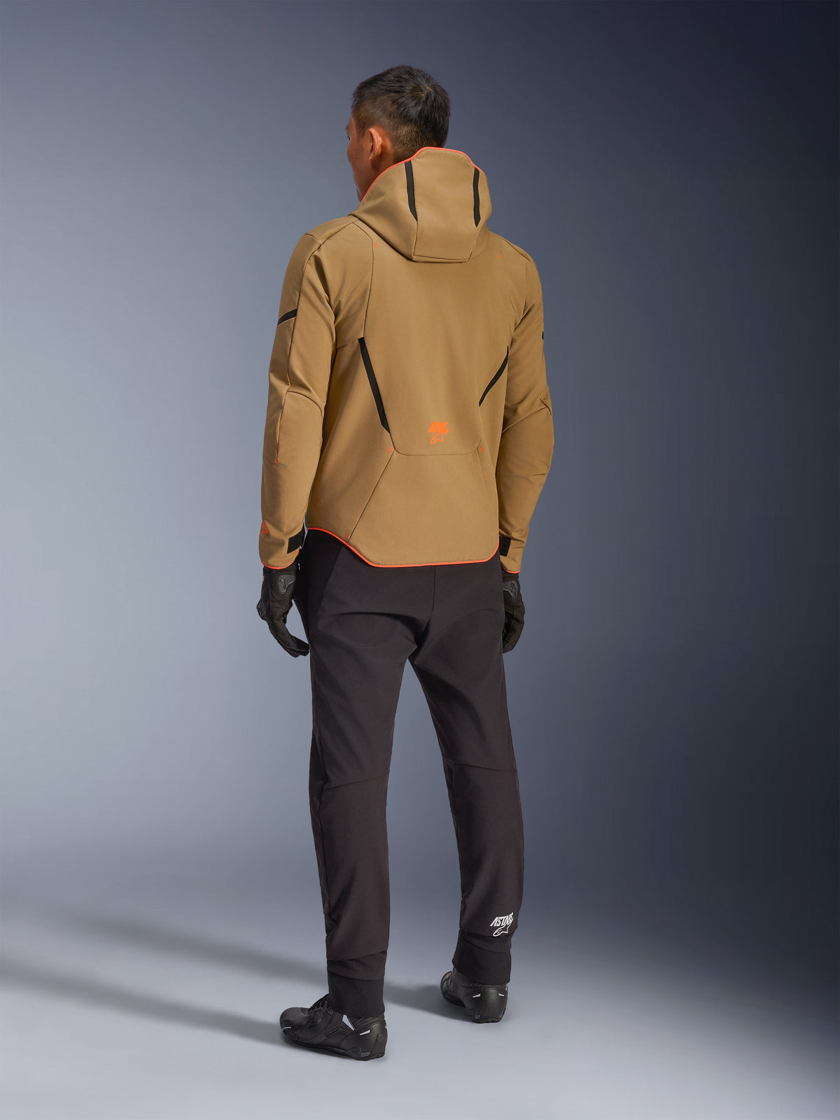 Alpinestars Aeroshell Waterproof Jacket, Dark Khaki Black Orange Fluo, vista posteriore di un pilota che indossa la giacca softshell con cappuccio, abbinata a pantaloni e guanti neri, con dettagli geometrici neri e un logo arancione sulla parte inferiore della schiena.