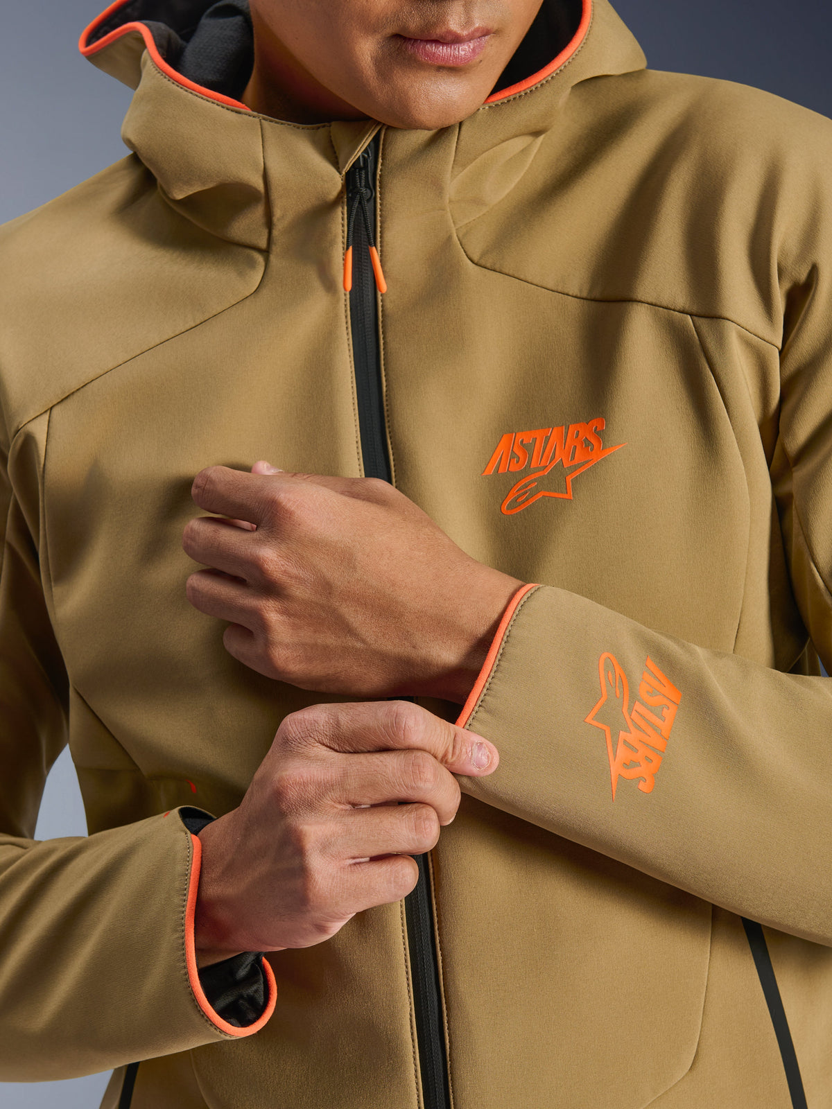 Giacca Alpinestars Aeroshell Waterproof, Dark Khaki e Arancione Fluo, primo piano della parte anteriore e della manica con loghi ASTARS arancioni, profili arancioni su cappuccio e polsini e cerniera impermeabile nera.
