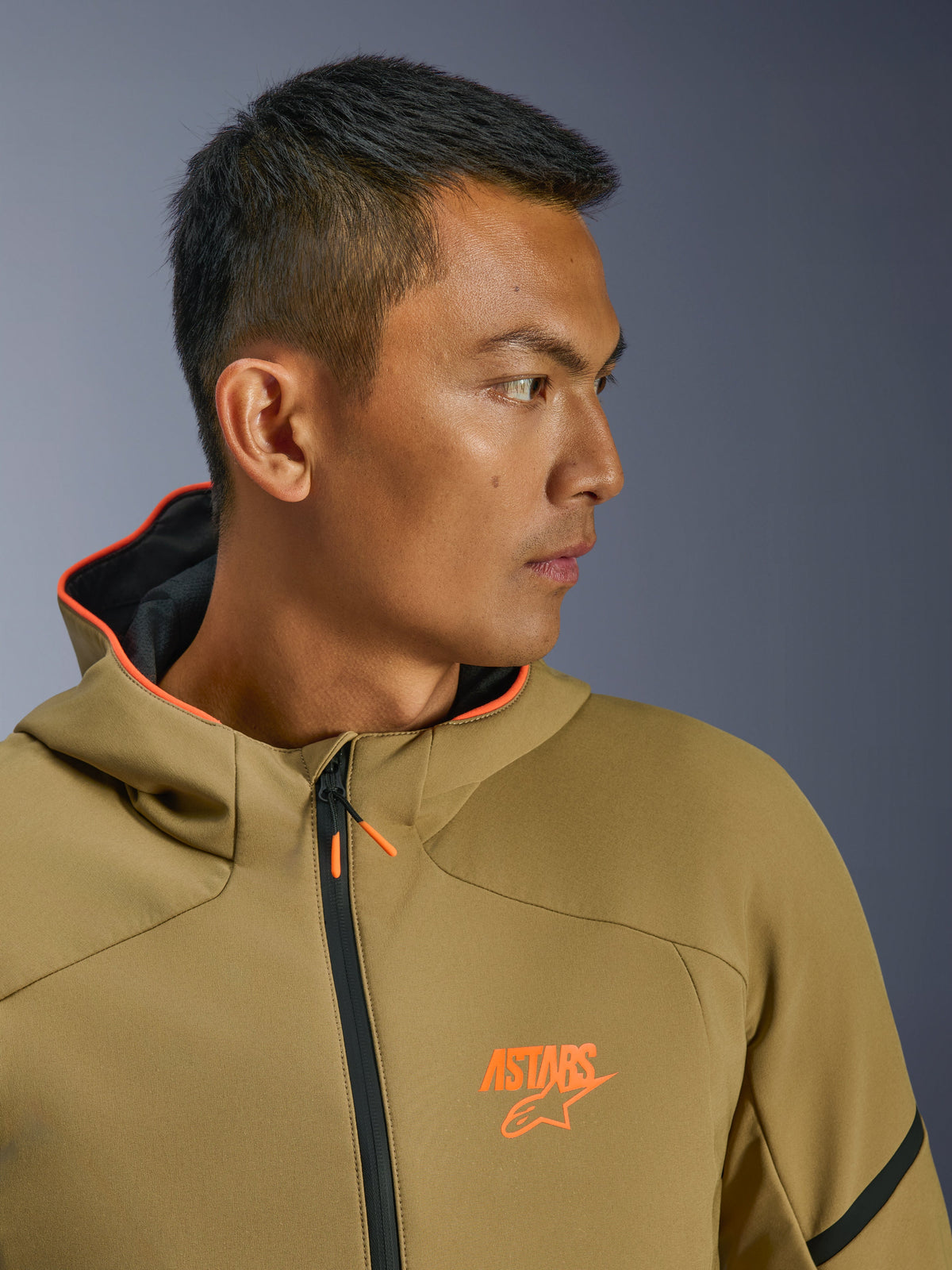 Giacca Alpinestars Aeroshell Waterproof, Dark Khaki e Arancione Fluo, profilo laterale ravvicinato, con logo ASTARS arancione sul petto e cappuccio con interno arancione, moderna felpa softshell crossover per la guida della moto