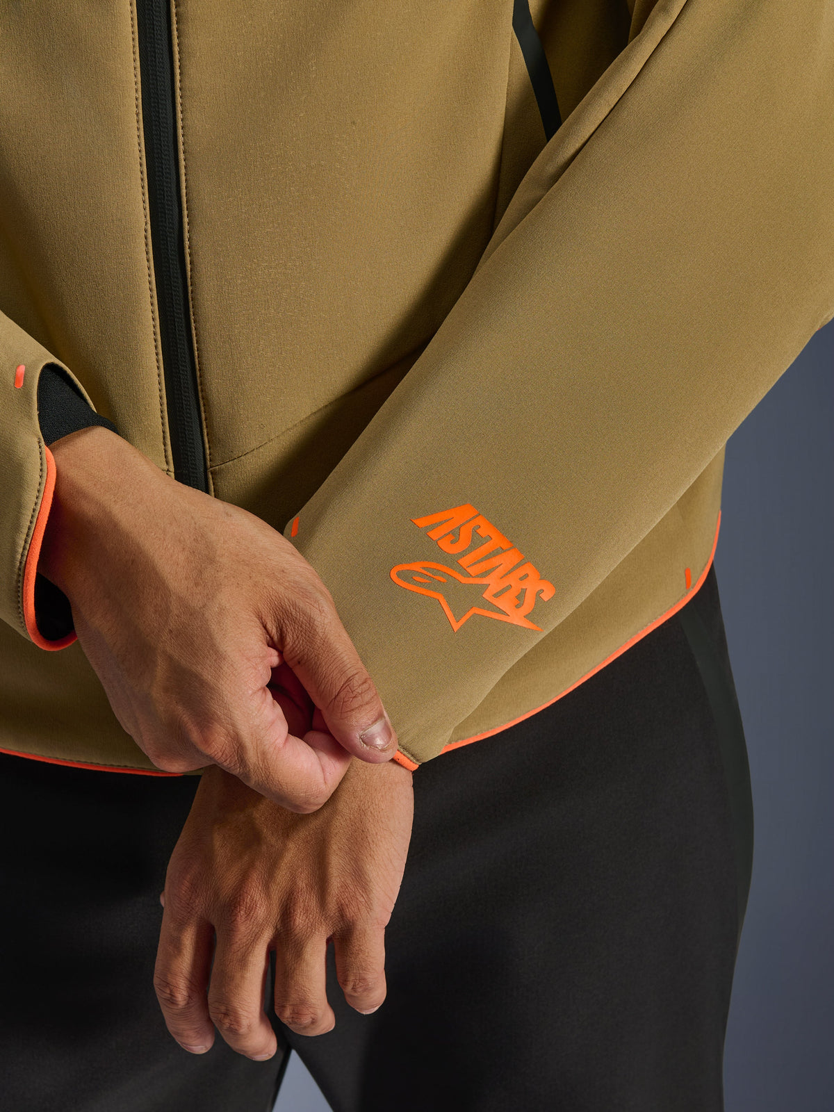 Giacca Alpinestars Aeroshell Waterproof, Dark Khaki con accenti Orange Fluo, primo piano del polsino e dell'orlo inferiore con logo Astars arancione brillante, profili arancione neon e dettaglio della cerniera frontale impermeabile, costruzione softshell sportiva.