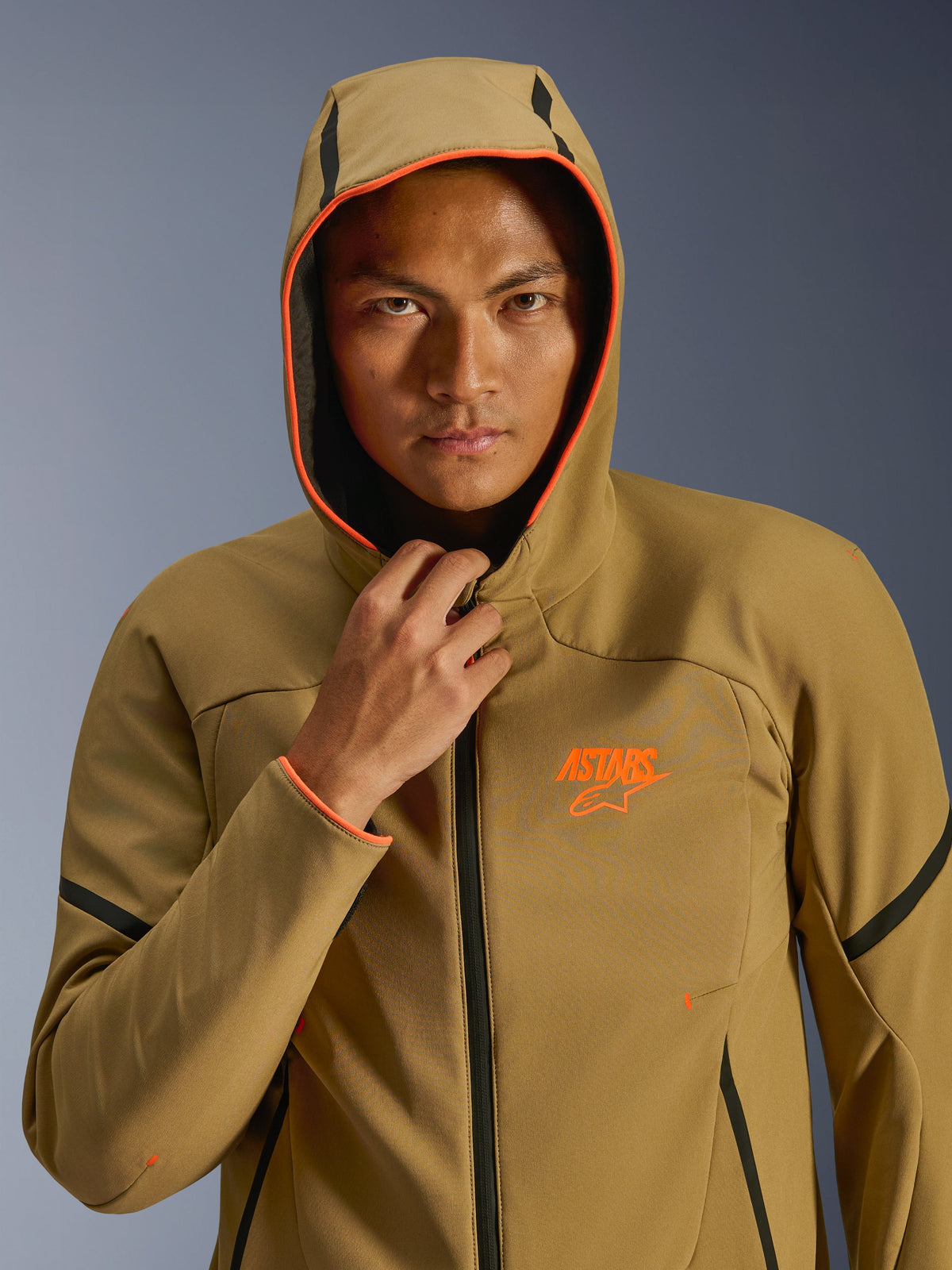 Giacca Alpinestars Aeroshell Waterproof, Dark Khaki, nero e arancione fluo, modello che indossa la giacca con il cappuccio alzato, caratterizzata da vivaci bordi arancione fluo e logo ASTARS arancione, eleganti dettagli neri, softshell impermeabile dal design sportivo moderno