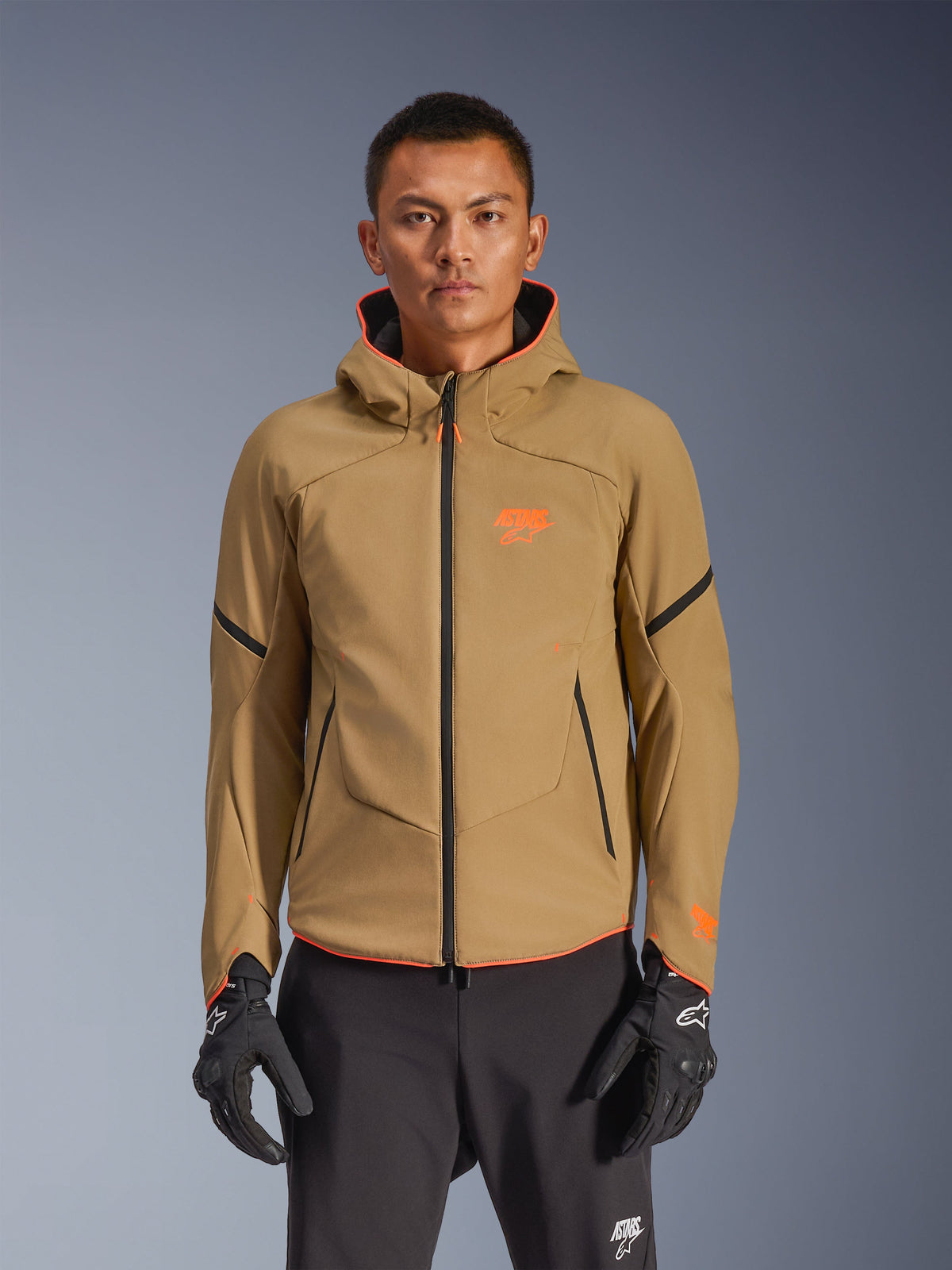 Alpinestars Aeroshell Waterproof Jacket, giacca da moto con cappuccio color kaki scuro con loghi arancioni e cerniere nere, linee di accento nere sulle maniche, indossata con guanti e pantaloni neri
