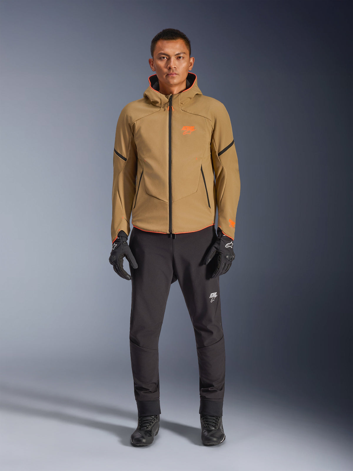 Modello che indossa la giacca Alpinestars Aeroshell Waterproof in Dark Khaki con accenti neri e arancione fluo, logo arancione sul petto e design con cappuccio, abbinata a pantaloni neri e guanti da moto neri, stile casual sport-touring