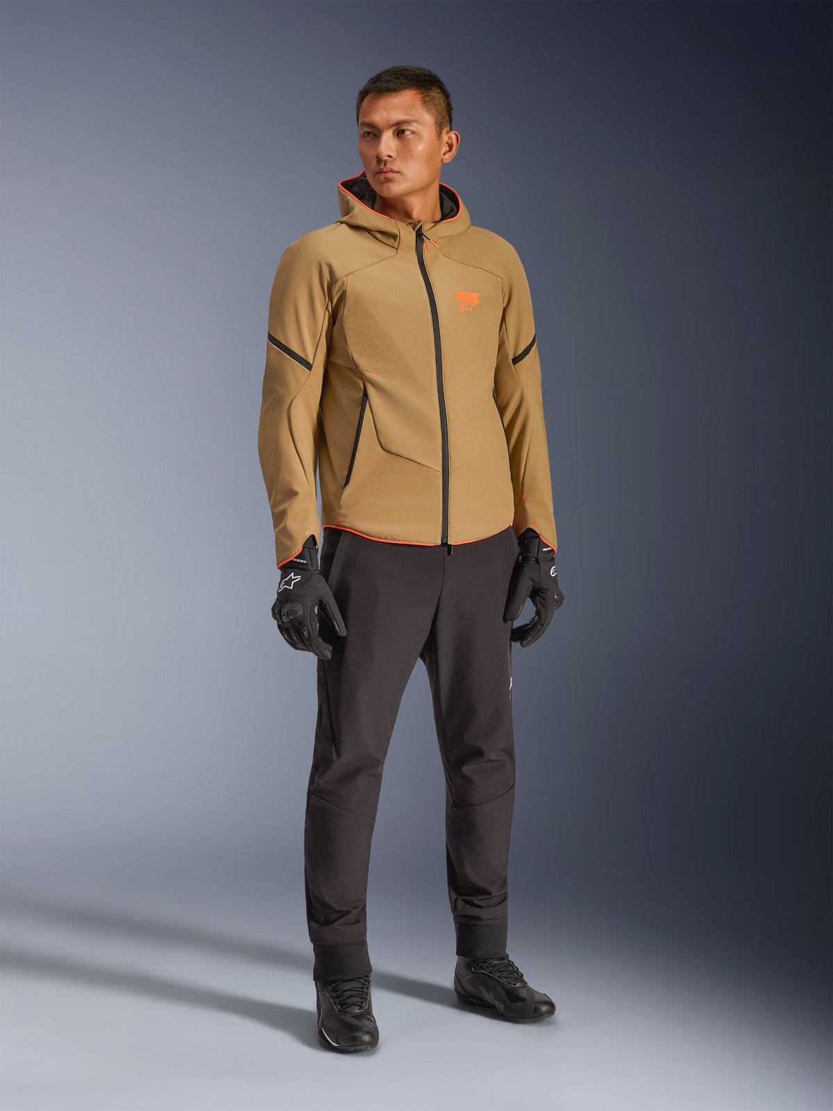 Giacca Alpinestars Aeroshell Waterproof in Dark Khaki, nero e arancione fluo, indossata da un modello con abbigliamento coordinato, caratterizzata da un design softshell con cappuccio, cerniere impermeabili e vivaci accenti e loghi arancioni.