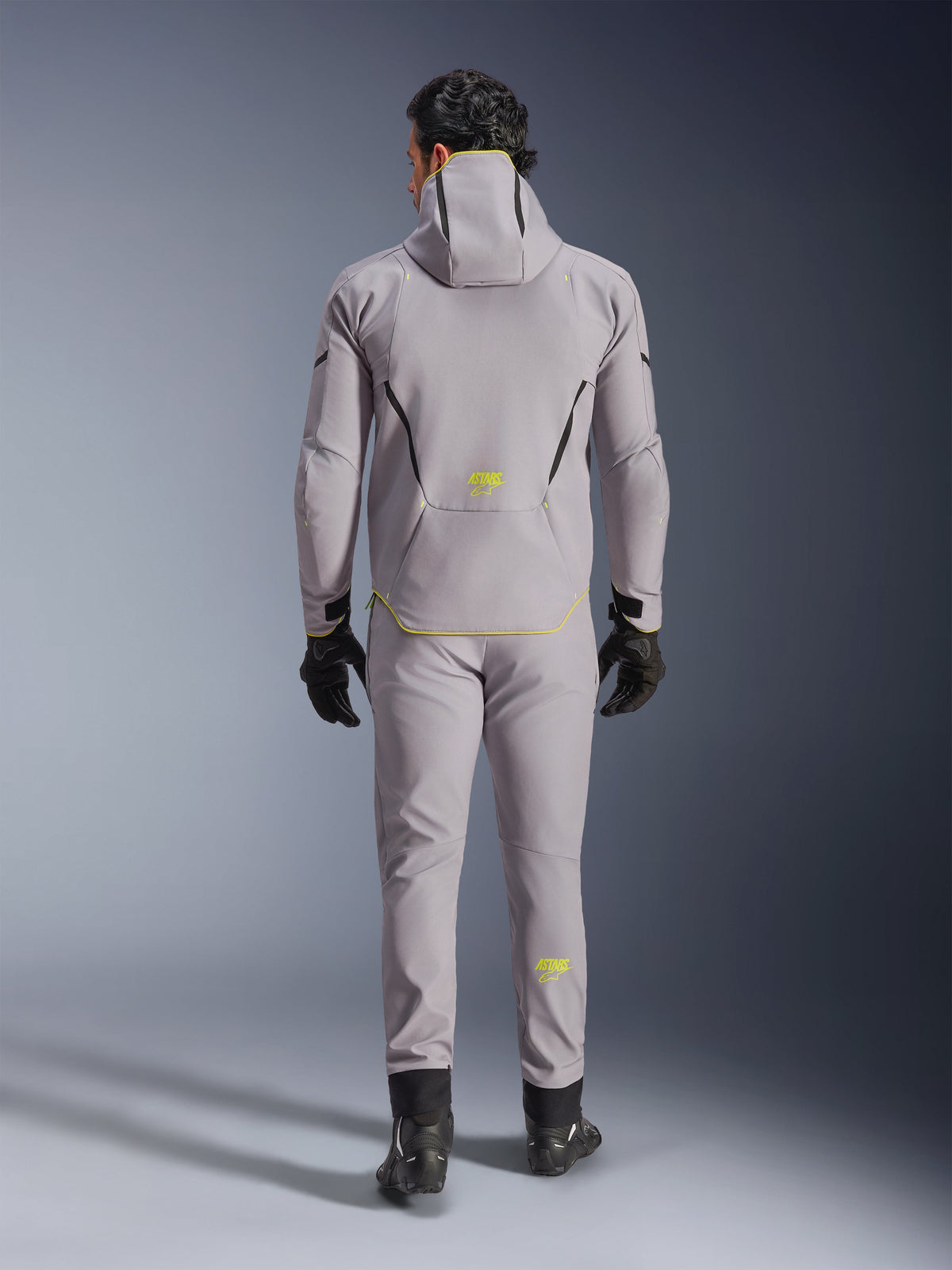 Giacca Alpinestars Aeroshell Waterproof in grigio medio e giallo fluo, vista posteriore completa, indossata con pantaloni grigi coordinati, guanti neri e scarpe da moto nere. Caratterizzata da un design softshell con cappuccio, marchio 'ASTARS' e dettagli giallo fluo, progettata per prestazioni stradali moderne.
