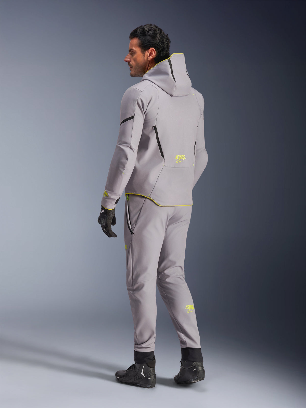 Giacca Alpinestars Aeroshell Waterproof, grigio medio e giallo fluo, indossata con pantaloni grigi coordinati, caratterizzata da un design con cappuccio con accenti e loghi gialli, vista posteriore, abbinata a guanti e stivali da moto neri, abbigliamento moderno da motociclista