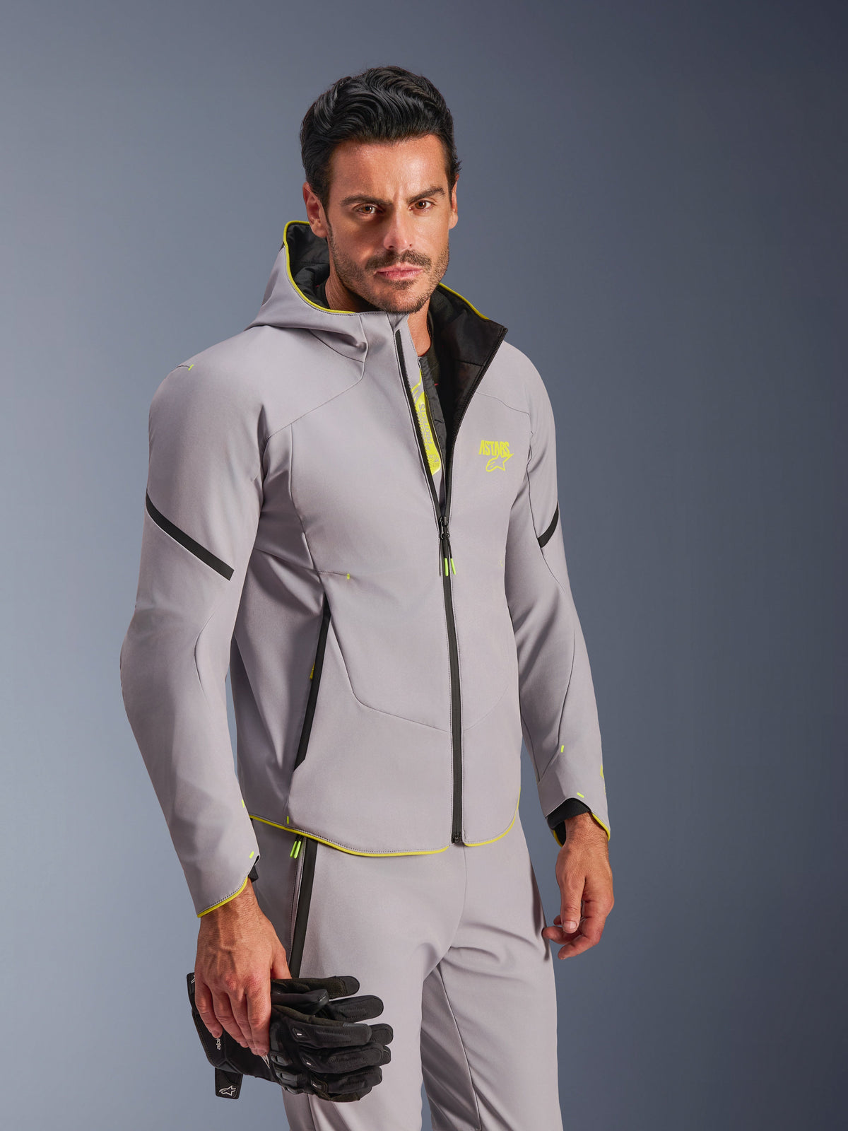 Giacca Alpinestars Aeroshell WP impermeabile in grigio medio e giallo fluo, indossata da un uomo con pantaloni grigi coordinati e guanti da moto neri, design softshell con cappuccio, accenti giallo neon e logo sul petto, moderno equipaggiamento sportivo da moto.