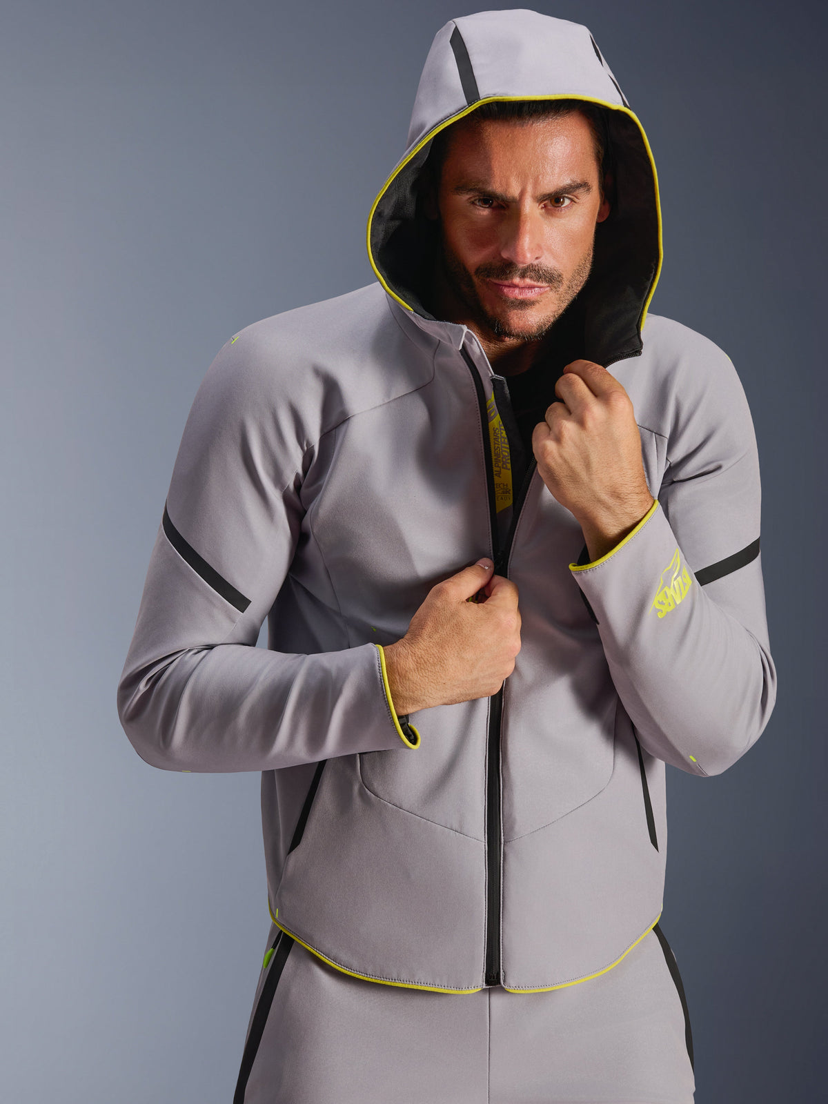 Giacca impermeabile Alpinestars Aeroshell WP, Grigio Medio e Giallo Fluo, giacca softshell sportiva con cappuccio, indossata da un modello con il cappuccio alzato, caratterizzata da cerniere impermeabili nere e dettagli giallo fluorescente su polsini e bordo inferiore, design moderno ispirato al motorsport