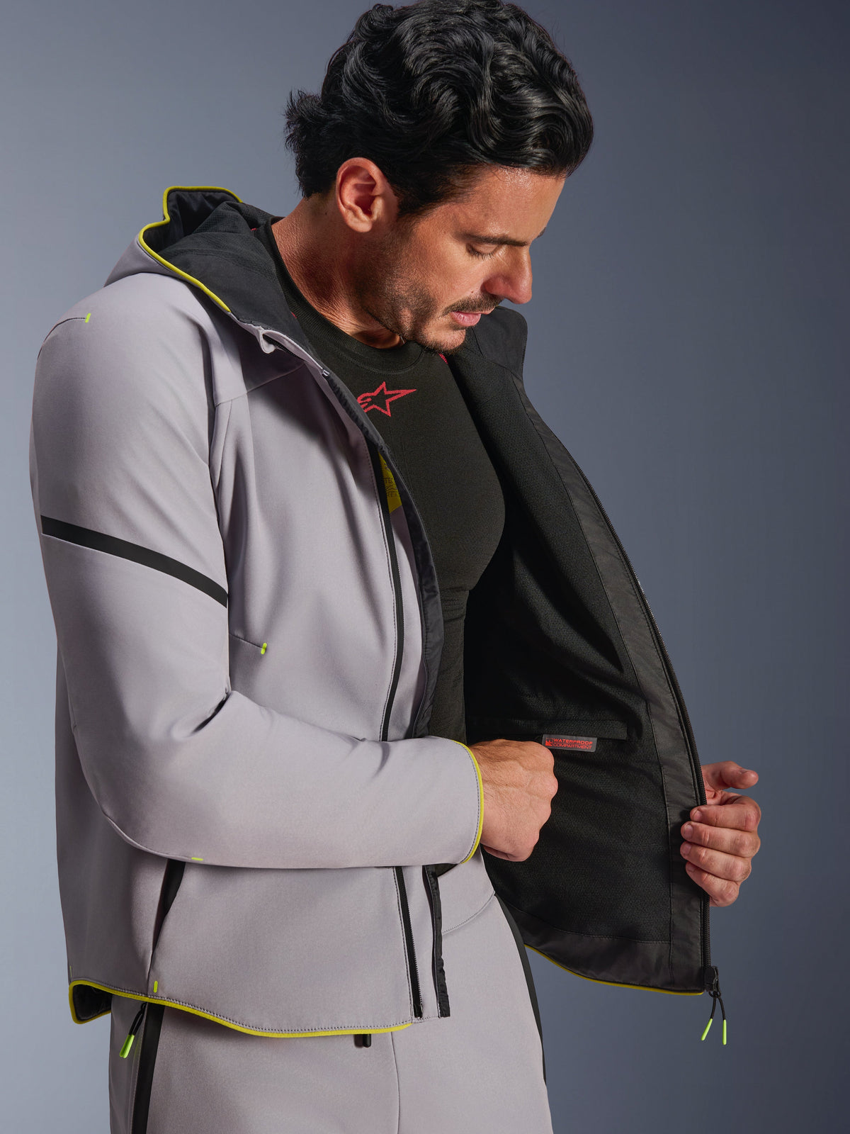Giacca Alpinestars Aeroshell Waterproof in Mid Gray e Yellow Fluo, indossata da un modello maschile che mostra lo scomparto interno impermeabile e la fodera nera, felpa sportiva da moto in softshell con cappuccio, finiture in giallo fluorescente e vestibilità atletica moderna.