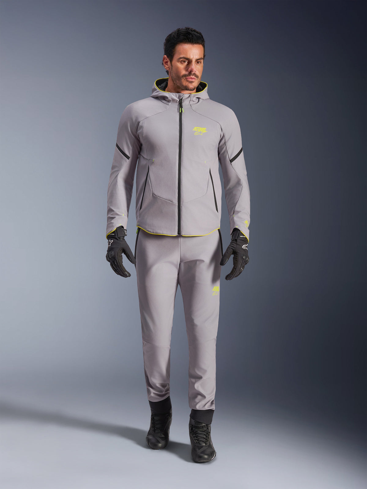 Giacca impermeabile Alpinestars Aeroshell, Mid Gray Yellow Fluo, indossata con pantaloni grigi coordinati, guanti e stivali neri, caratterizzata da un moderno design a felpa con cappuccio di ispirazione sportiva con vivaci accenti gialli e vestibilità atletica.