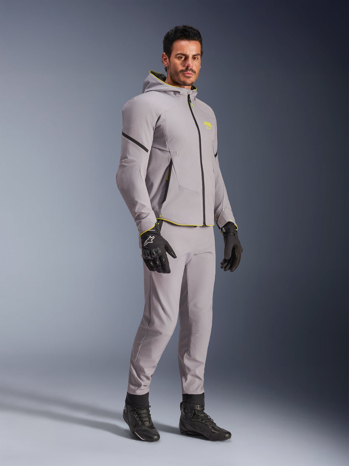 Giacca Alpinestars Aeroshell Waterproof, grigio medio e giallo fluo, indossata da un modello con pantaloni grigi coordinati, guanti da moto neri e stivali neri, abbigliamento da moto moderno in stile felpa con cappuccio per guida casual e sportiva.
