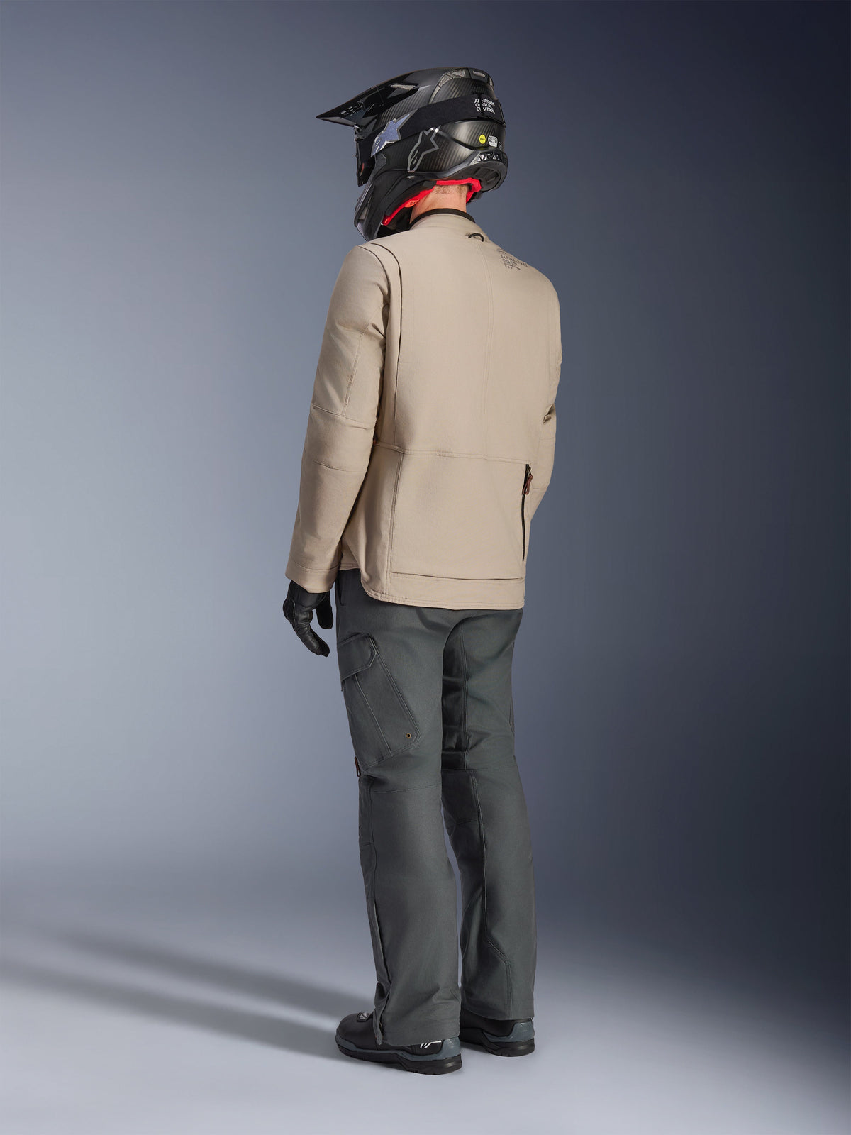 Giacca Alpinestars Borrego Drystar®, giacca da moto adventure Warm Gray, vista posteriore del pilota che indossa pantaloni grigio scuro e un casco nero, caratterizzata da un look robusto in tela cerata e un design funzionale.