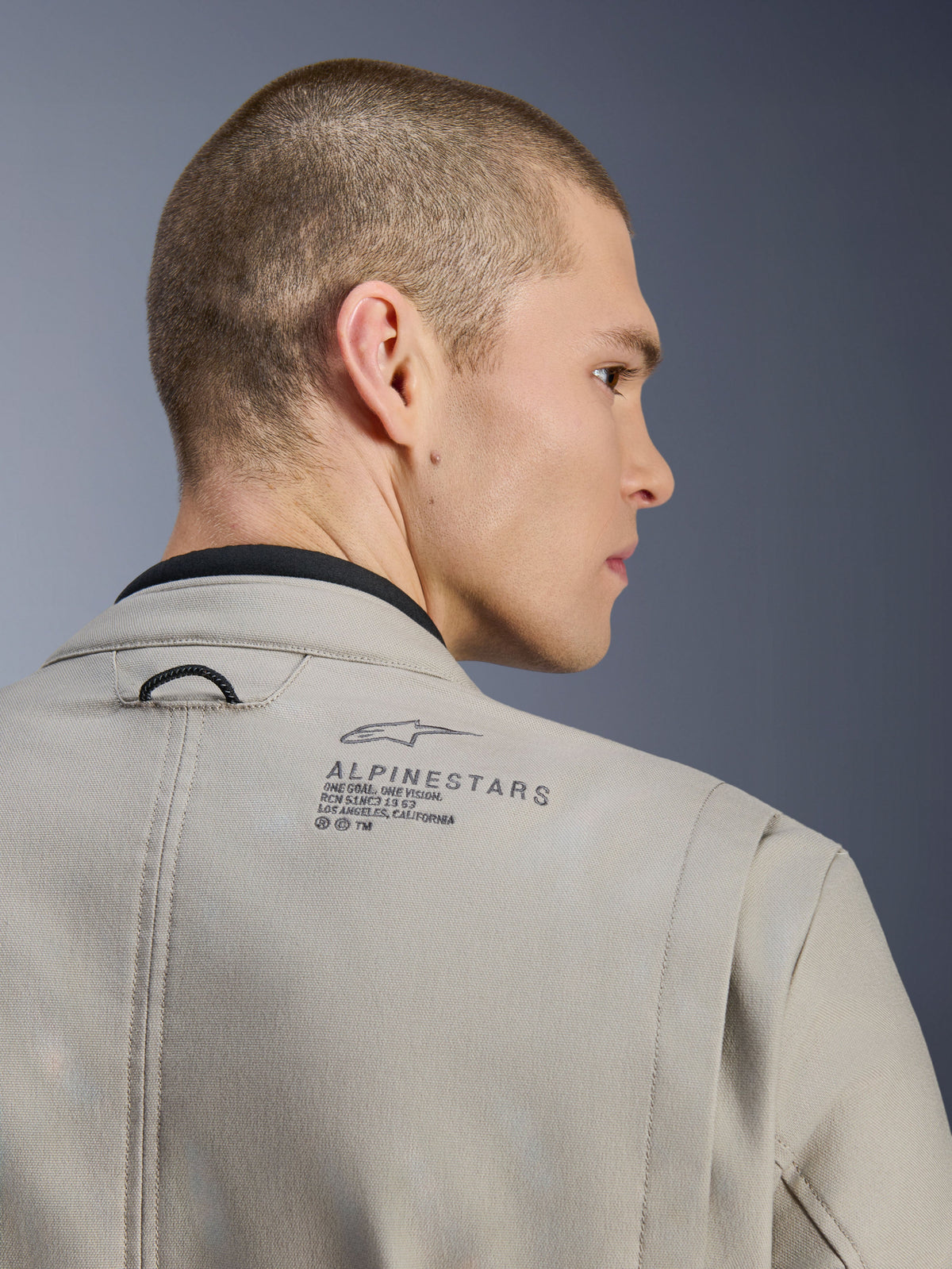 Giacca Alpinestars Borrego Drystar® in Warm Gray, primo piano della spalla posteriore, materiale in tela cerata testurizzata, logo Alpinestars nero e testo heritage, indossata da un modello, giacca da moto adventure