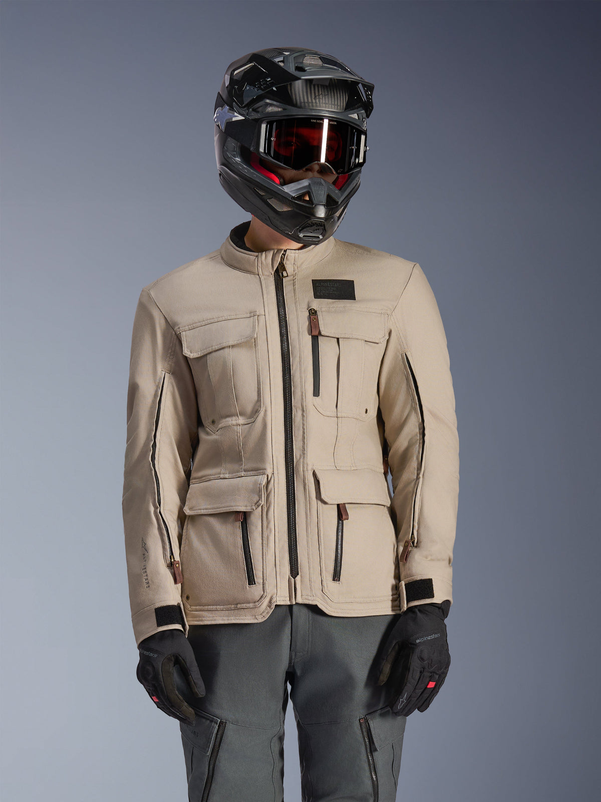 Giacca Alpinestars Borrego Drystar®, giacca da moto adventure, grigio caldo, con tasche cargo frontali multiple e cerniere nere, indossata con casco nero, occhiali e pantaloni da moto grigio scuro, design in tessuto robusto per il touring