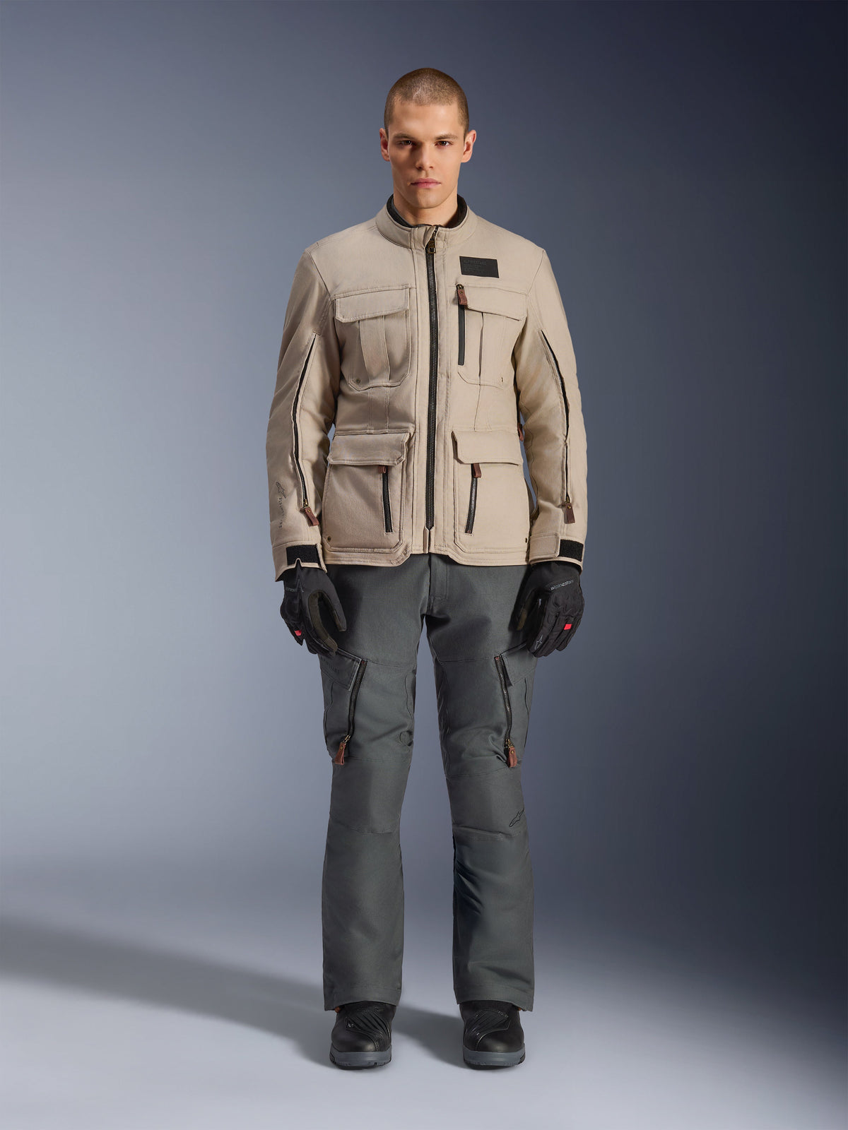 Giacca Alpinestars Borrego Drystar®, giacca da moto adventure, grigio Warm Gray, indossata da un modello con pantaloni da guida grigio scuro e guanti neri, con numerose tasche cargo e una robusta costruzione in tessuto dal classico look in tela cerata
