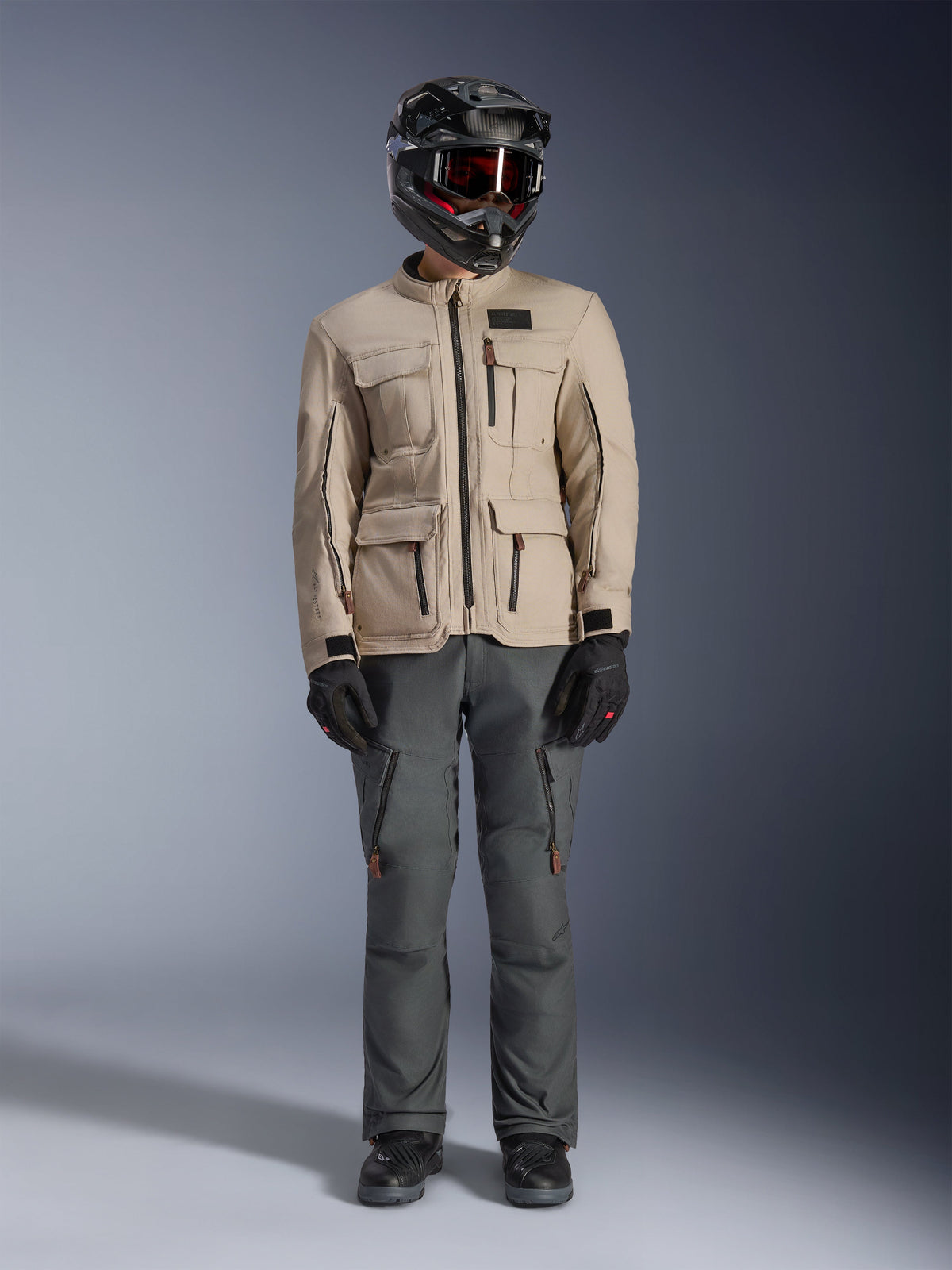 Giacca Alpinestars Borrego Drystar® in Warm Gray, giacca da moto adventure con quattro tasche cargo anteriori, abbinata a pantaloni da guida grigio scuro, casco off-road nero e guanti neri, caratterizzata da un robusto look in tela cerata