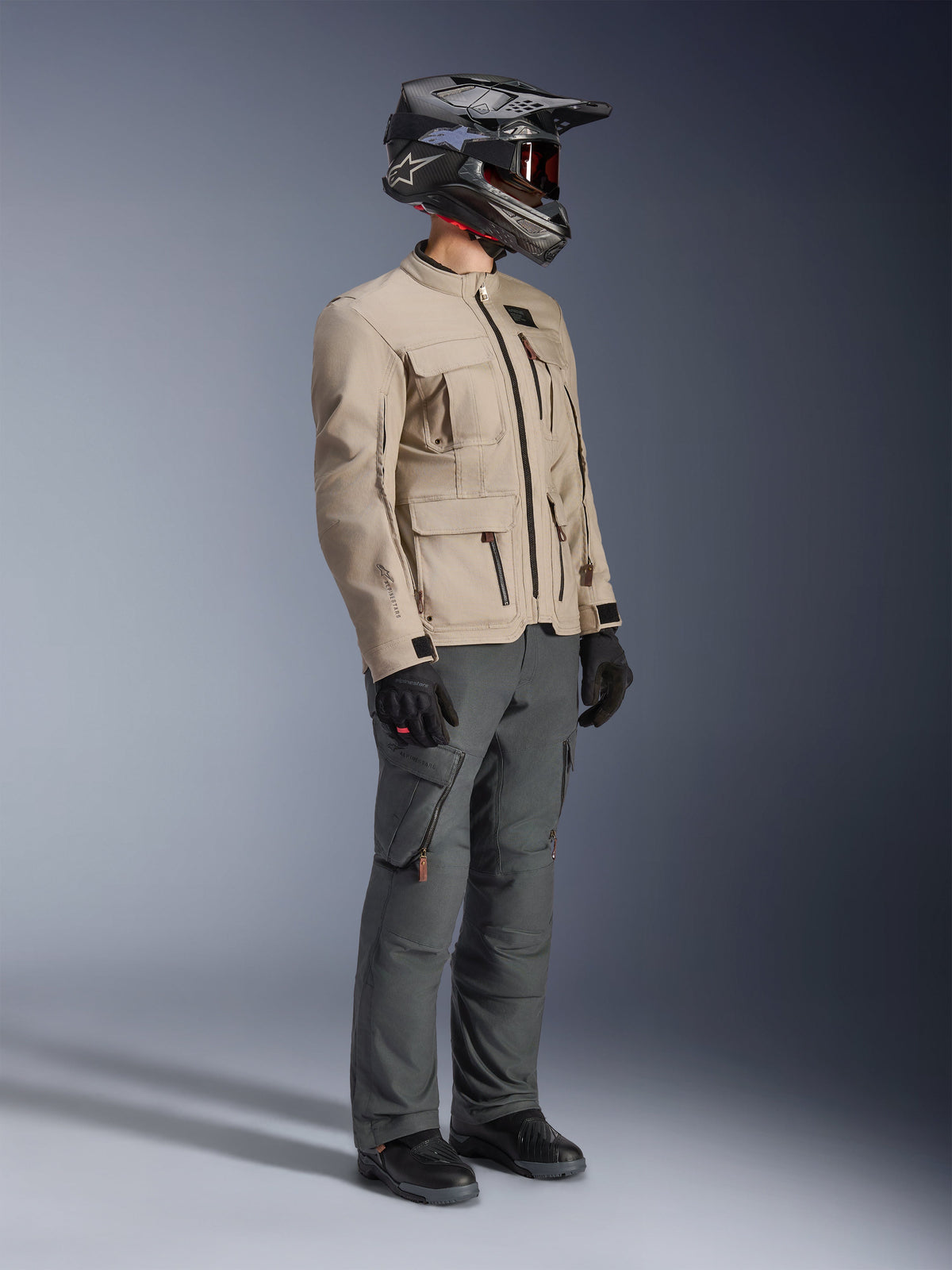 Motociclista che indossa la giacca Alpinestars Borrego Drystar® in Warm Gray, giacca da moto adventure con trama in tela cerata e tasche utility, abbinata a pantaloni da guida grigio scuro, casco in fibra di carbonio, guanti neri e stivali neri, vista a corpo intero in studio di un outfit completo da touring adventure.