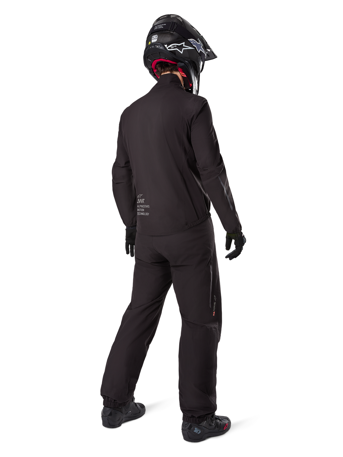 Vista posteriore di un pilota che indossa la fodera della giacca Alpinestars AMT-8 Stretch Drystar® XF, abbigliamento tecnico da moto nero, abbinato a pantaloni coordinati e casco nero, con il marchio "AMT Alpinestars Motion Technology" sul retro, design minimalista per l'adventure touring