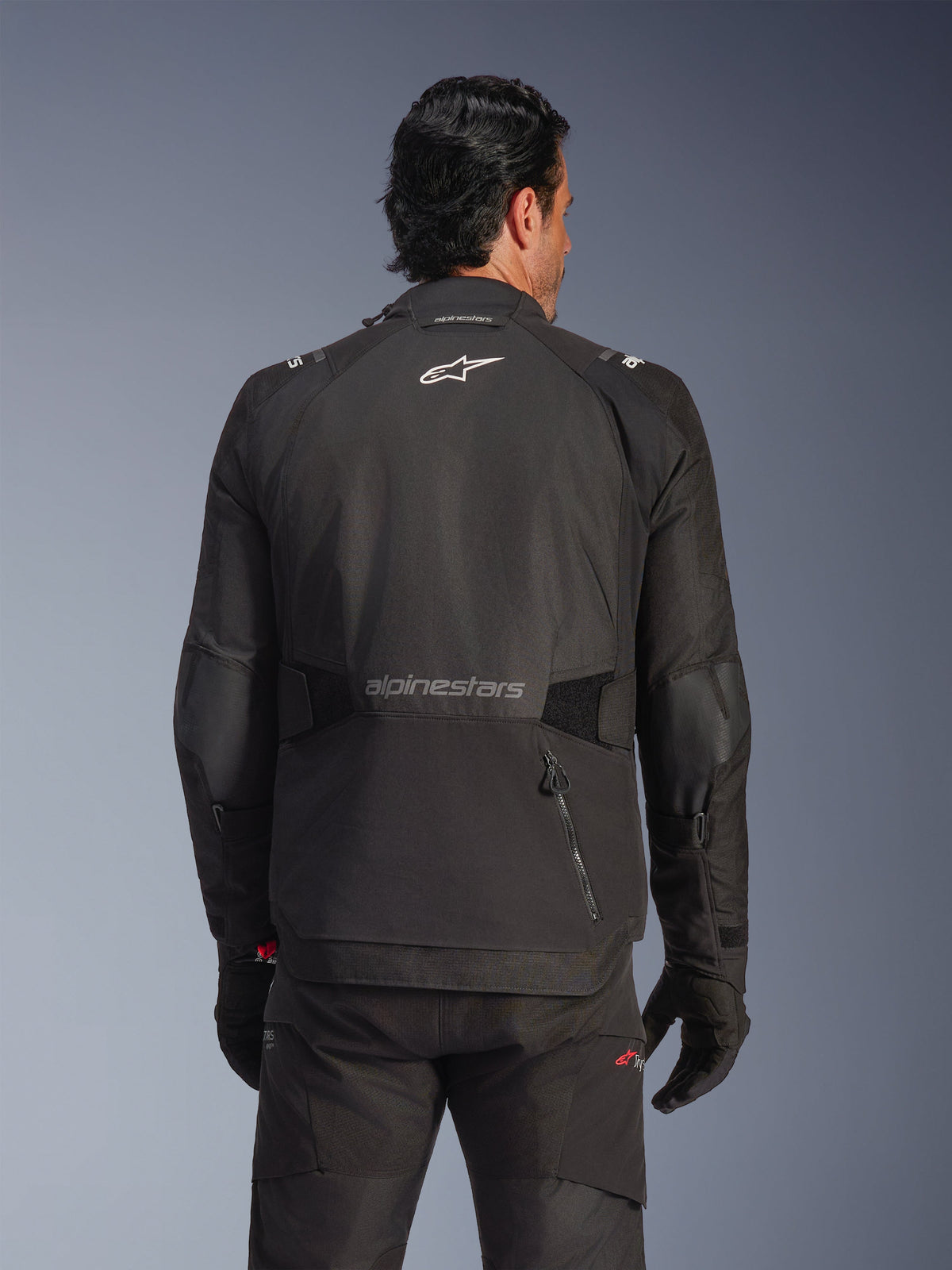 Giacca Alpinestars Andes V4 Drystar®, nero, vista posteriore su un modello, giacca adventure in tessuto con loghi riflettenti e tasca posteriore con zip, abbinata a pantaloni e guanti neri coordinati