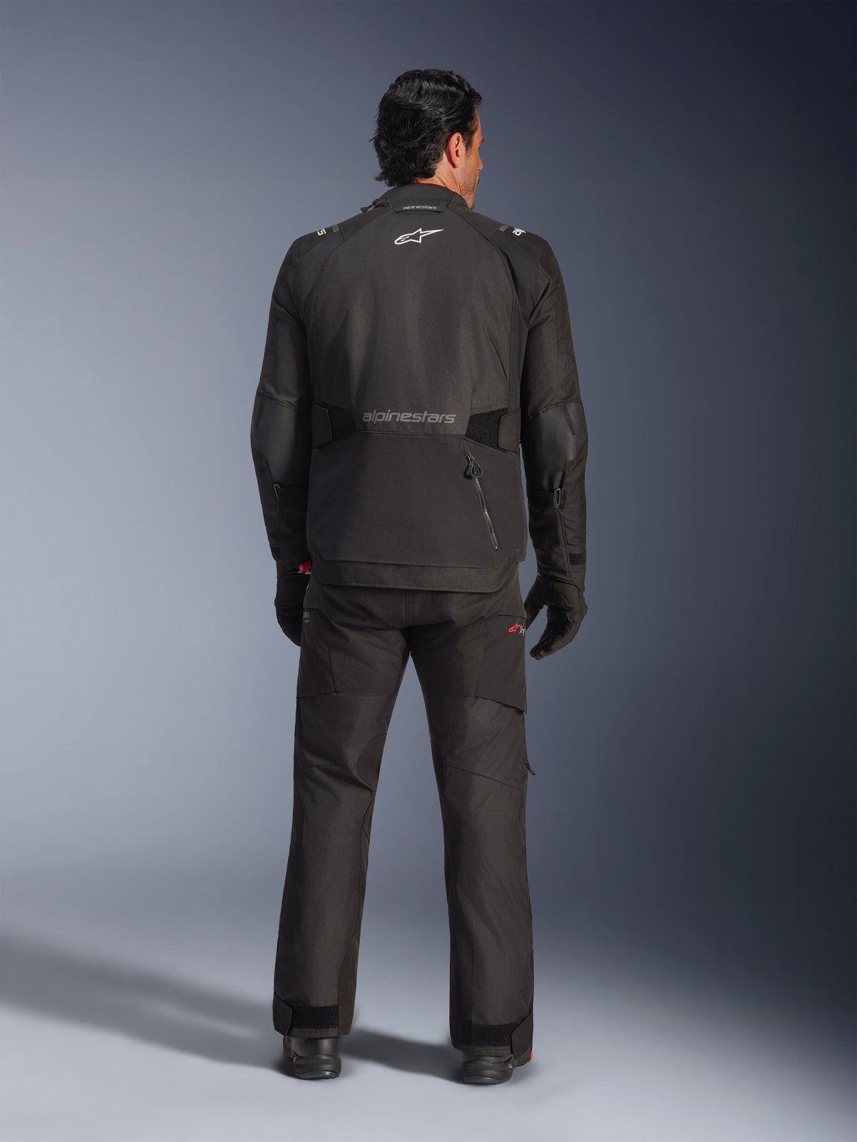 Vista posteriore di un pilota che indossa la giacca Alpinestars Andes V4 Drystar® in nero, abbinata a pantaloni e guanti da moto coordinati, con logo e scritta Alpinestars sulla schiena, abbigliamento tecnico adventure per ogni condizione atmosferica