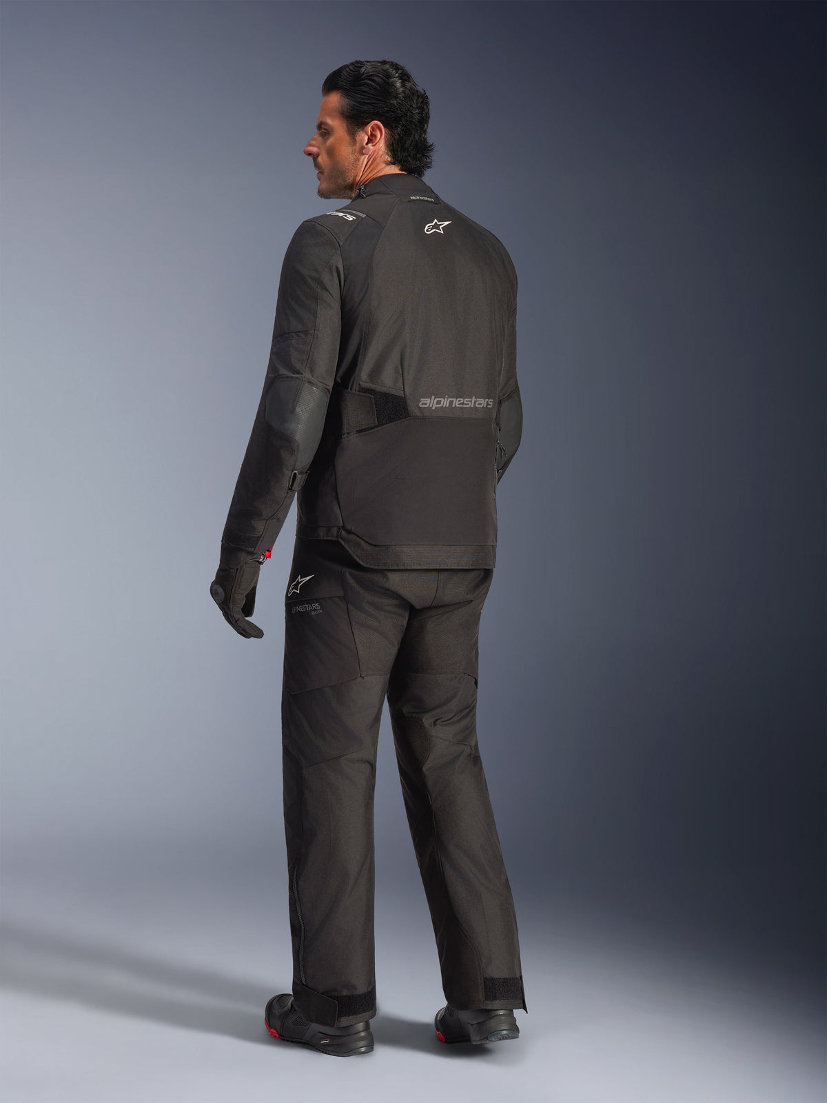 Giacca Alpinestars Andes V4 Drystar®, nero, vista posteriore del pilota in equipaggiamento completo da moto, giacca adventure in tessuto con marchio Alpinestars sulla schiena, indossata con pantaloni e stivali neri coordinati, stile road e adventure per ogni condizione atmosferica