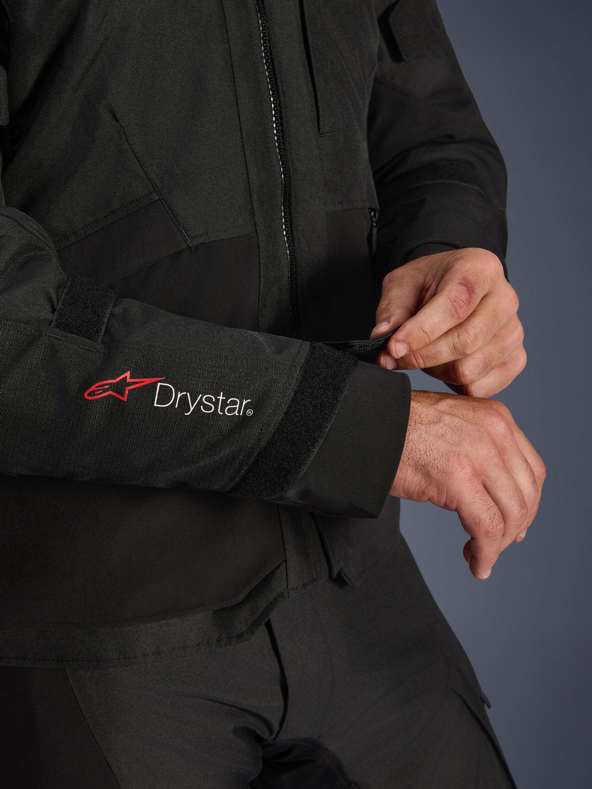 Giacca Alpinestars Andes V4 Drystar®, giacca da moto in tessuto nero, primo piano della manica con logo Alpinestars rosso e branding Drystar® bianco, pilota che regola il cinturino in velcro sul polso per una vestibilità sicura, dettaglio funzionale per la guida adventure