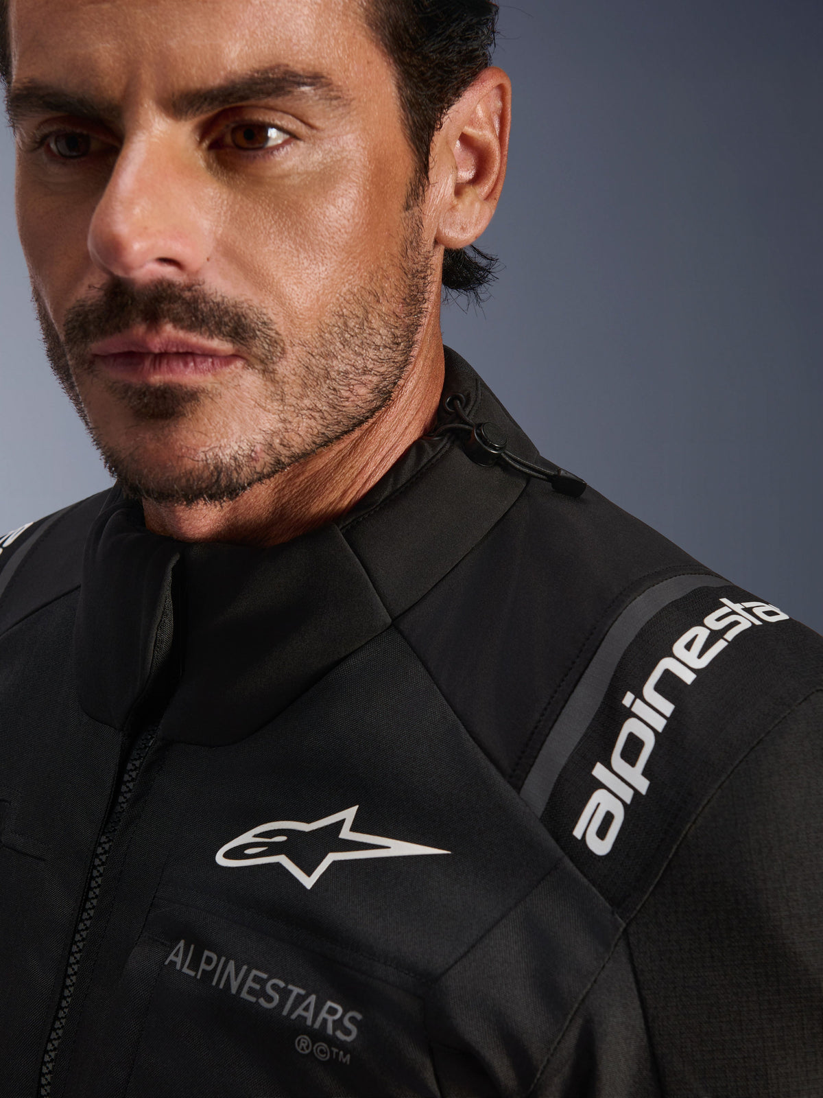 Giacca Alpinestars Andes V4 Drystar®, giacca da moto in tessuto nero, dettaglio ravvicinato indossato da un uomo, con loghi Alpinestars bianchi su petto e spalla, cordoncino del colletto regolabile e design adventure-touring ad alte prestazioni.