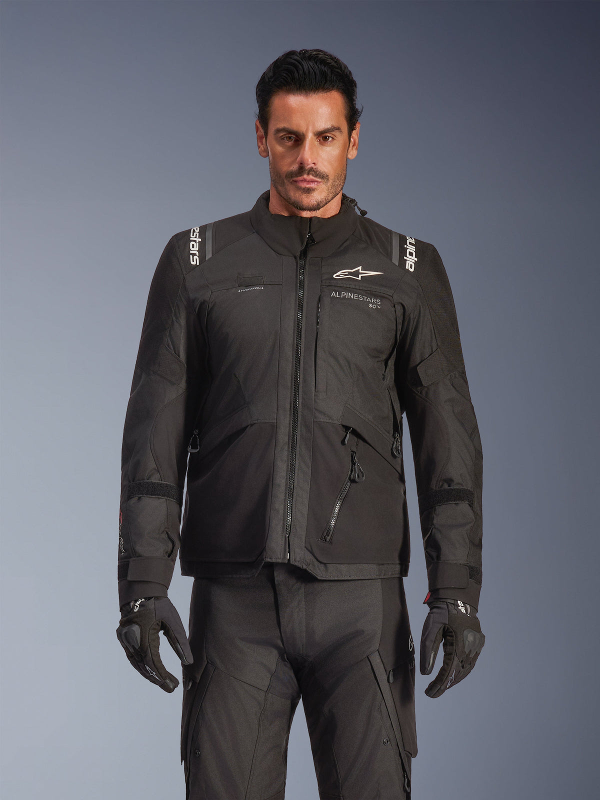 Modello maschile con giacca Alpinestars Andes V4 Drystar® nera, giacca da moto adventure touring, abbinata a pantaloni e guanti neri coordinati, con cerniere impermeabili e logo Alpinestars bianco su petto e spalle, scatto in studio per abbigliamento tecnico da moto per ogni condizione atmosferica.