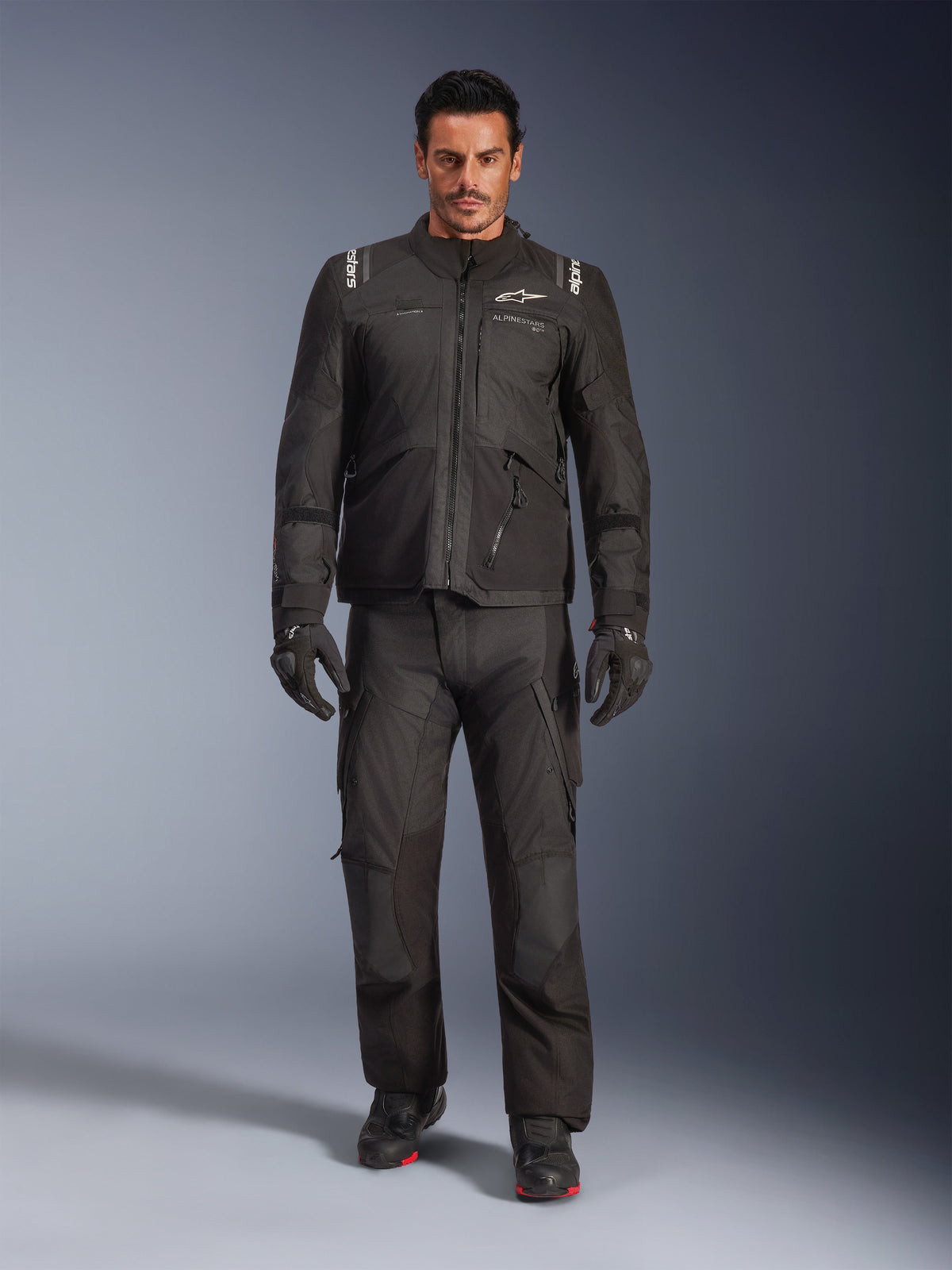 Modello che indossa la giacca Alpinestars Andes V4 Drystar® in nero, abbinata a pantaloni da moto adventure coordinati, guanti neri e stivali neri, scatto in studio a figura intera che mostra l'equipaggiamento tecnico da touring per tutte le stagioni con marchio Alpinestars.