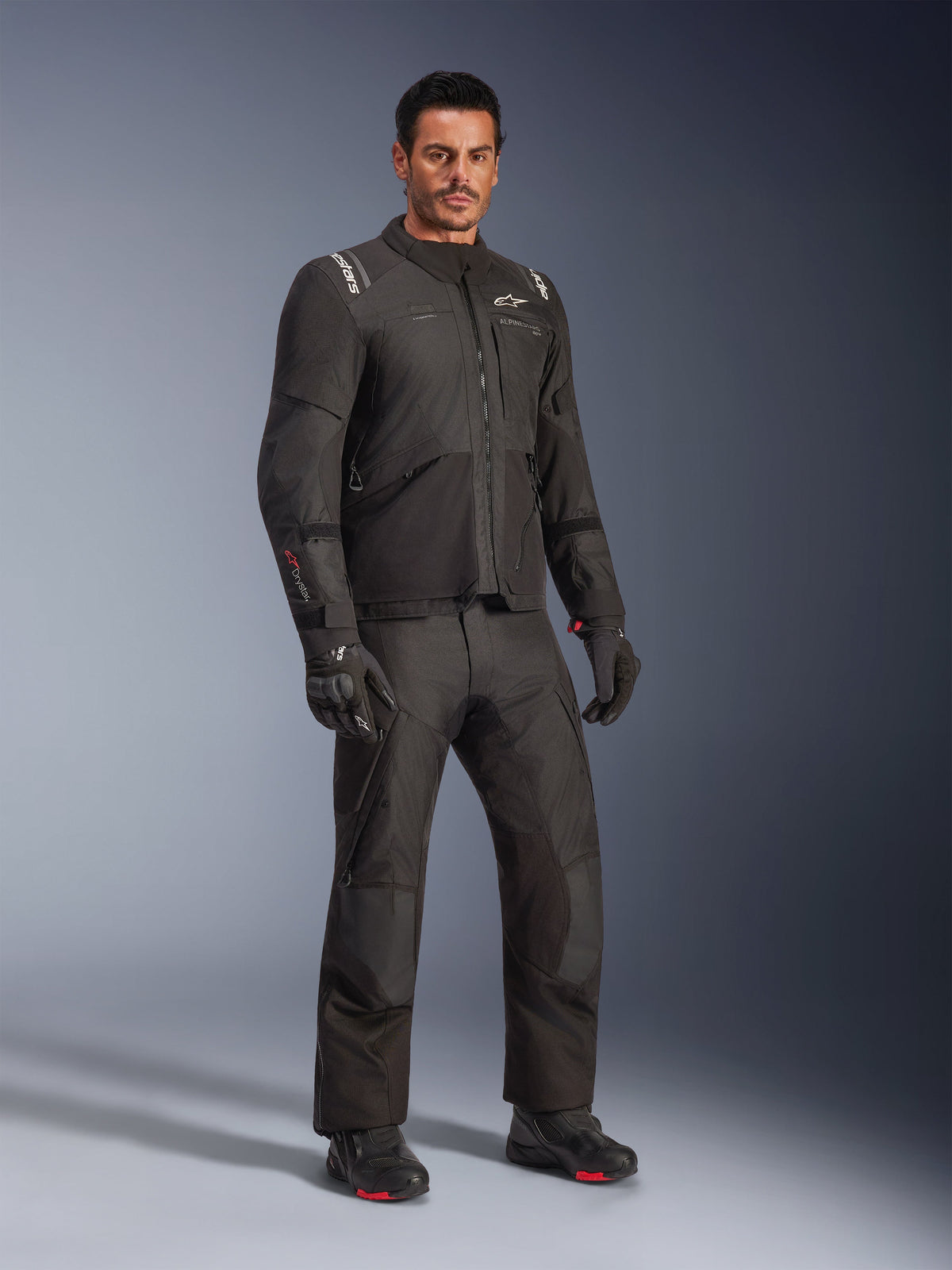 Giacca Alpinestars Andes V4 Drystar®, completo da moto nero indossato da un modello, giacca e pantaloni in tessuto adventure-touring, guanti e stivali neri, scatto in studio, design moderno per la guida in ogni condizione meteorologica