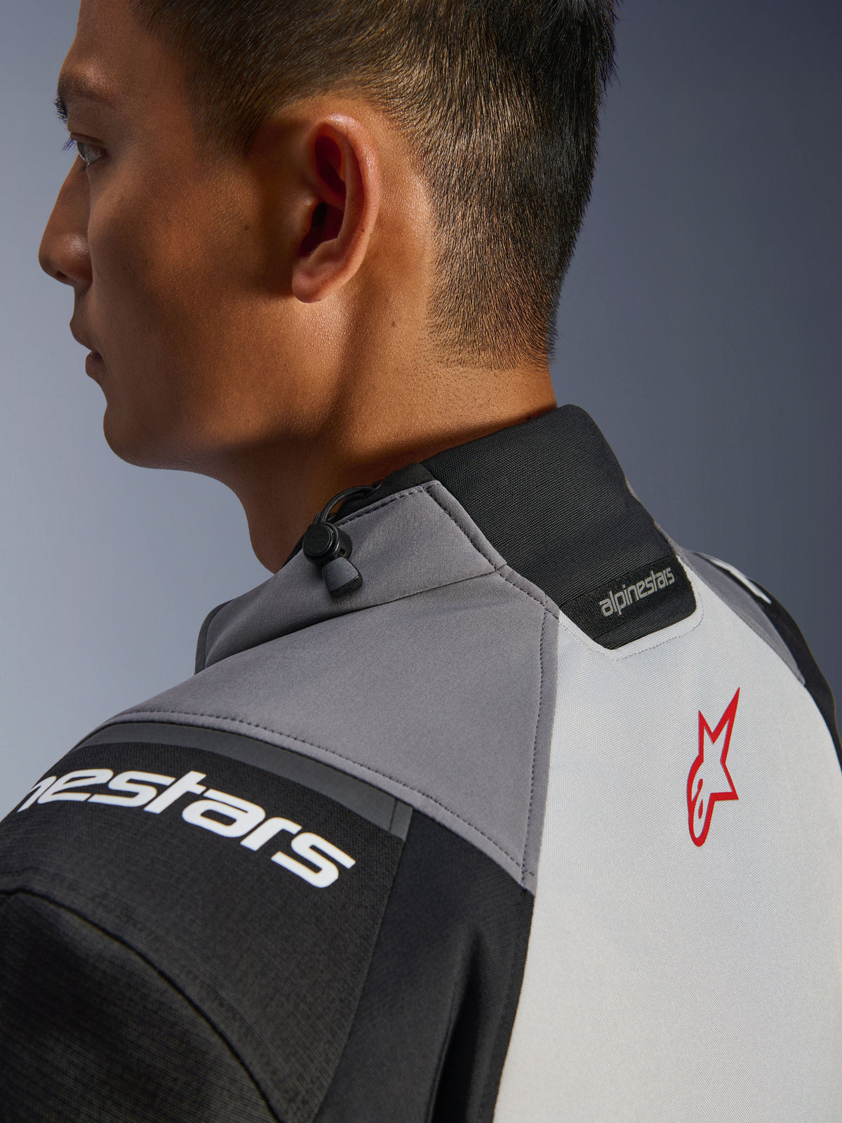 Giacca Alpinestars Andes V4 Drystar®, primo piano del colletto e della parte superiore della schiena, grigio scuro e grigio ghiaccio con accenti neri, logo a stella rosso, con regolatore a cordoncino sul colletto e marchio Alpinestars, struttura in tessuto resistente