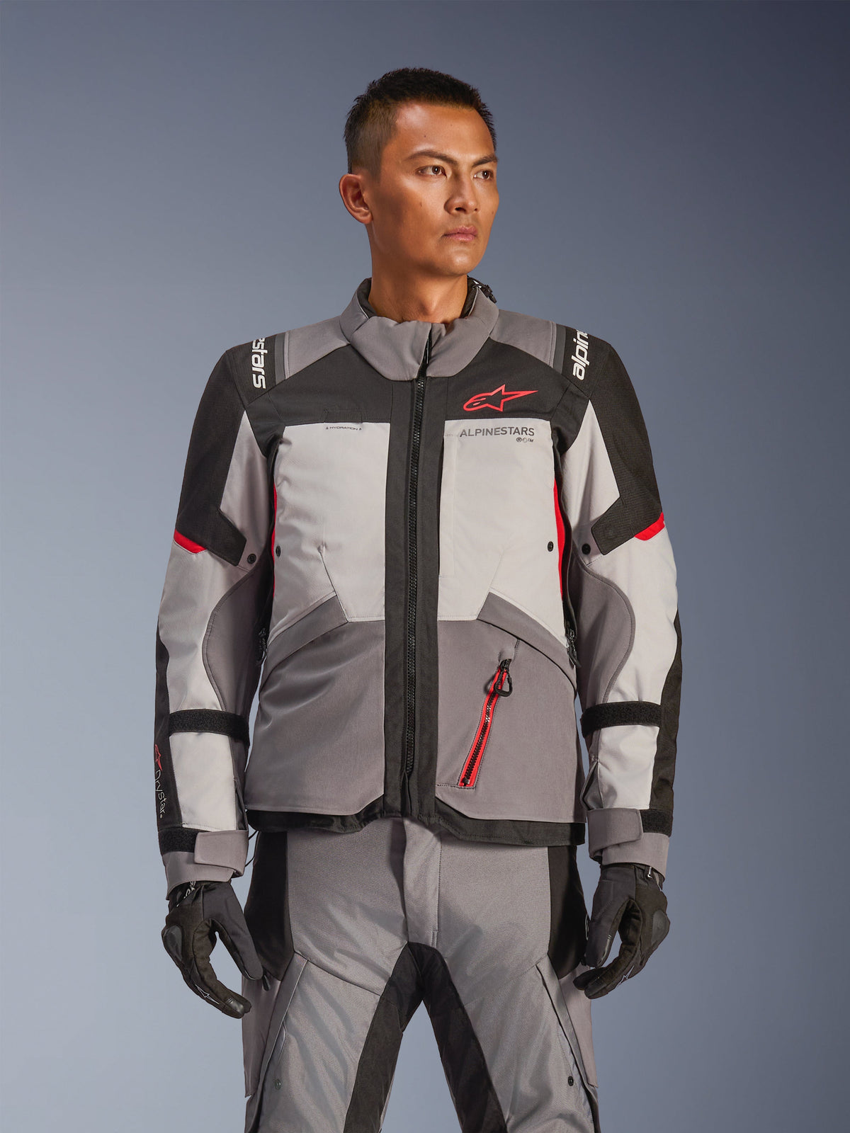 Giacca Alpinestars Andes V4 Drystar®, giacca da moto adventure touring, grigio scuro e grigio ghiaccio con dettagli neri e rosso acceso, indossata con pantaloni e guanti abbinati, design impermeabile e traspirante per la guida adventure in ogni condizione climatica