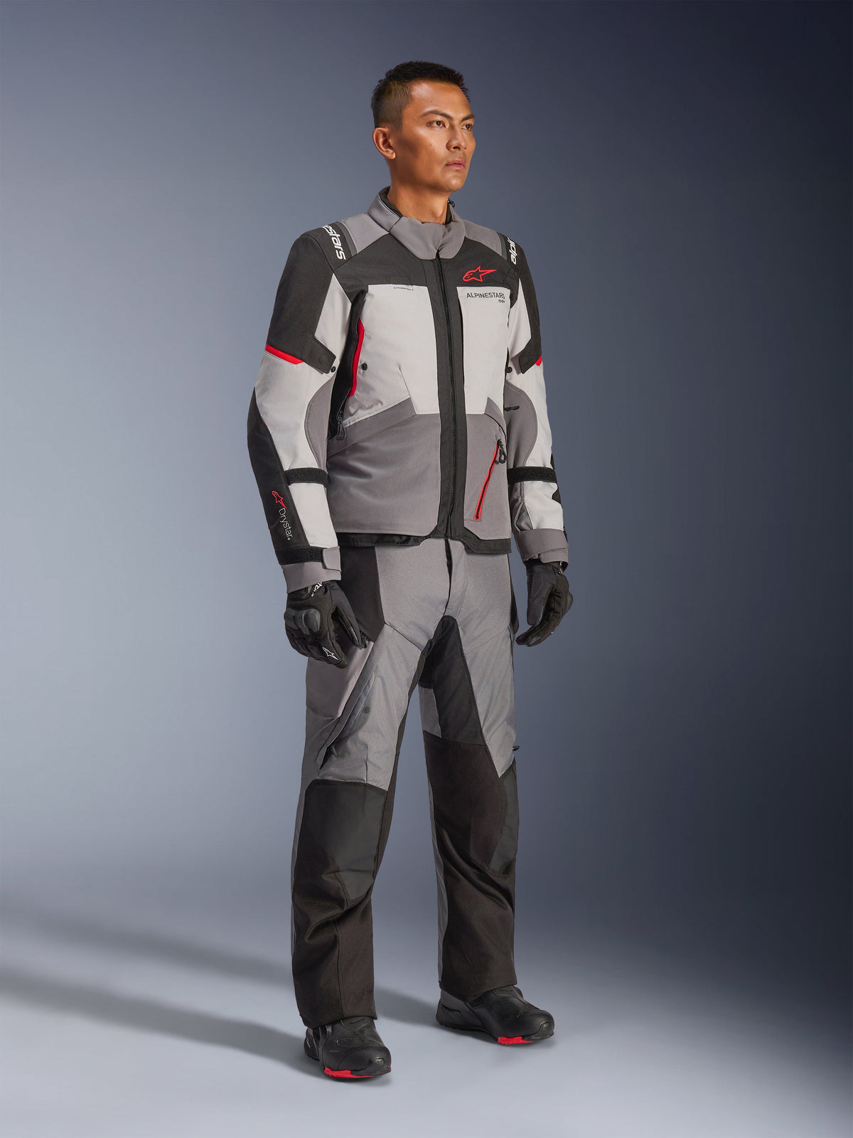 Modello con giacca Alpinestars Andes V4 Drystar® e pantaloni adventure coordinati, grigio scuro, grigio ghiaccio, nero e rosso acceso, abbinati a guanti e stivali da moto neri, abbigliamento tecnico per ogni condizione climatica con design moderno a più pannelli.