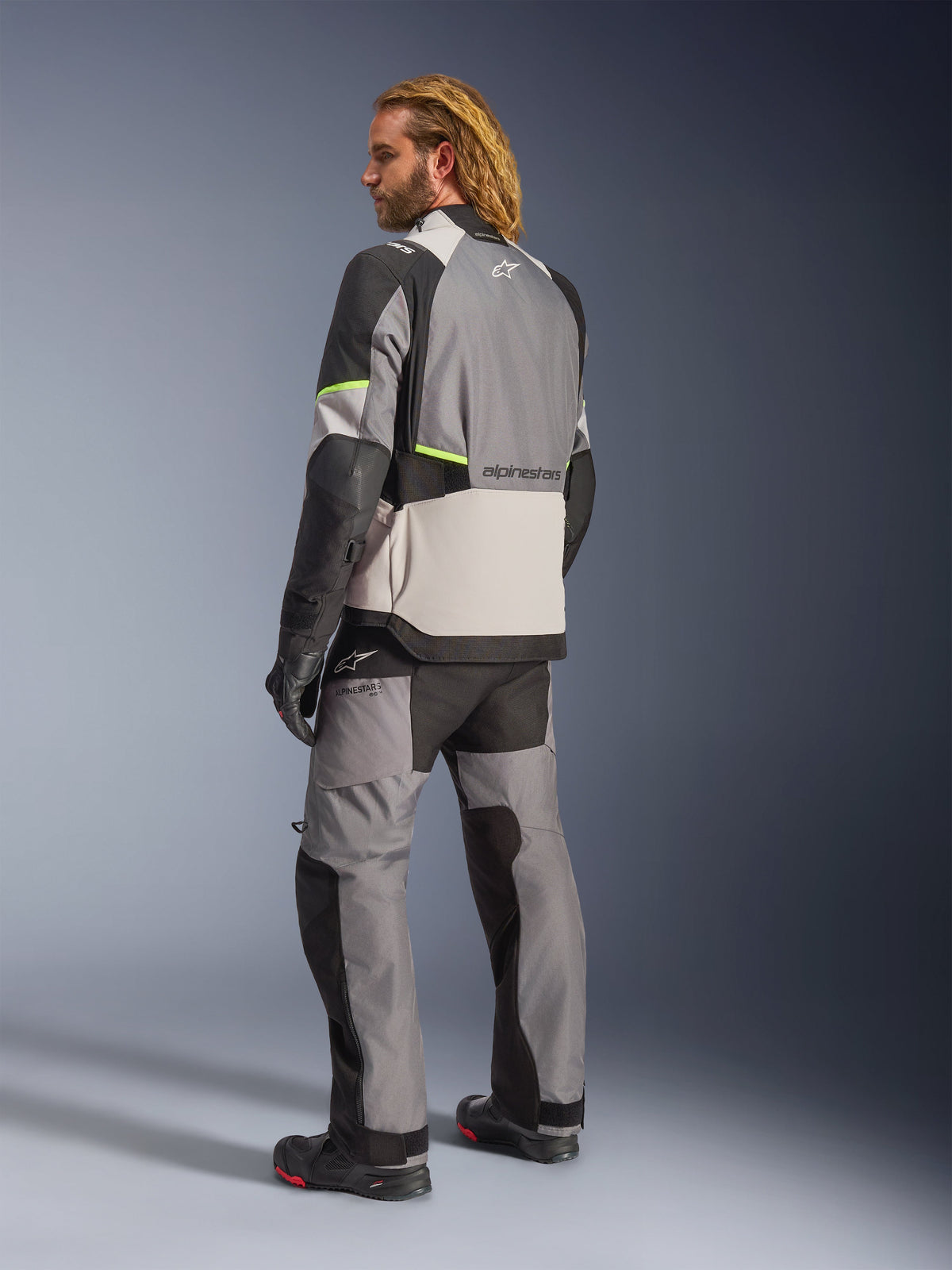 Giacca Alpinestars Andes V4 Drystar®, grigio scuro, grigio ghiaccio, nero e giallo fluo, vista posteriore del pilota che indossa pantaloni e stivali adventure touring coordinati, pannelli in tessuto grigio rinforzato, accenti giallo fluo e marchio Alpinestars, abbigliamento da moto per tutte le stagioni