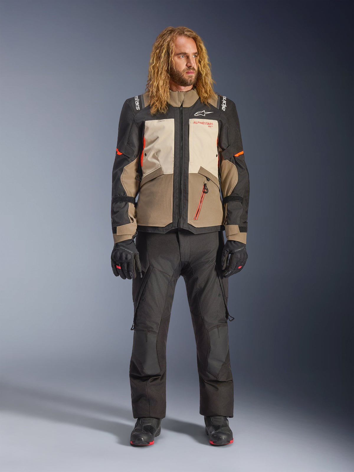 Giacca Alpinestars Andes V4 Drystar®, Walnut Mastic Black, indossata con pantaloni e guanti da moto neri, design adventure multi-pannello con accenti arancioni e marchio Alpinestars, abbigliamento tecnico in tessuto per ogni condizione atmosferica