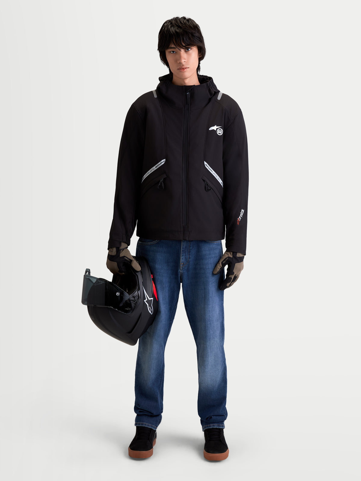 Alpinestars C 5 Drystar Giacca, giacca da moto urbana contemporanea da uomo in nero, vista frontale su un modello che indossa jeans e guanti e tiene in mano un casco, con costruzione softshell impermeabile e cappuccio integrato