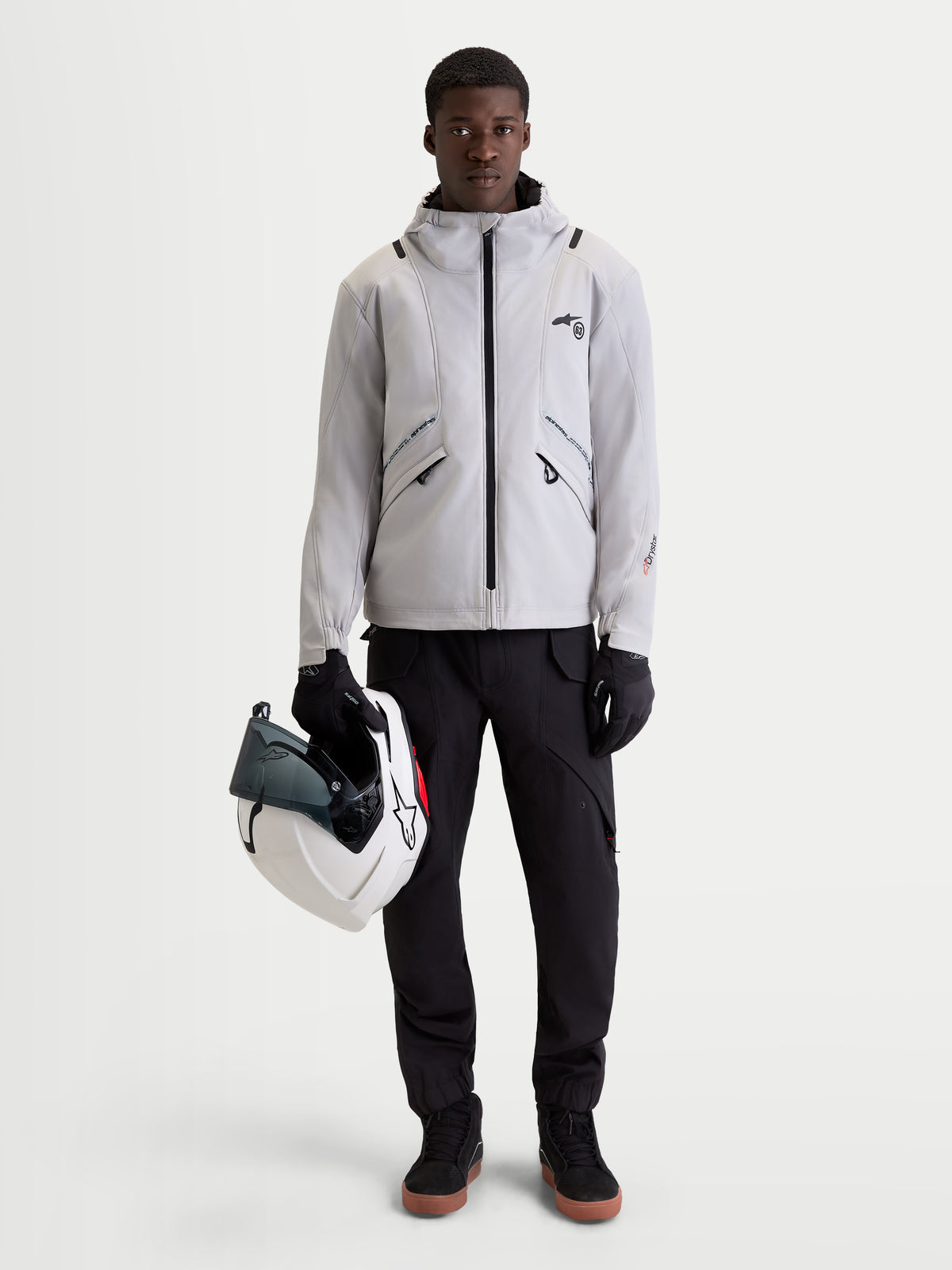 Alpinestars C 5 Drystar Giacca, giacca da moto urbana contemporanea da uomo in grigio chiaro e nero, vista frontale di un modello che indossa la giacca impermeabile con cappuccio integrato e tiene in mano un casco