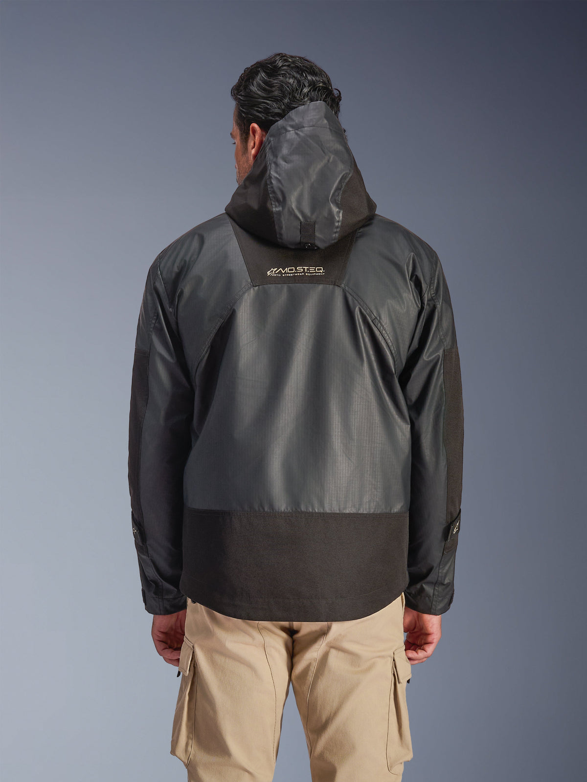 Vista posteriore di un uomo che indossa la giacca impermeabile Alpinestars Mospher in nero, con cappuccio e struttura in tessuto ripstop resistente, mostrata con pantaloni cargo beige, evidenziando il logo MO.ST.EQ. sulla parte superiore della schiena, design urban dell'attrezzatura da motociclista.